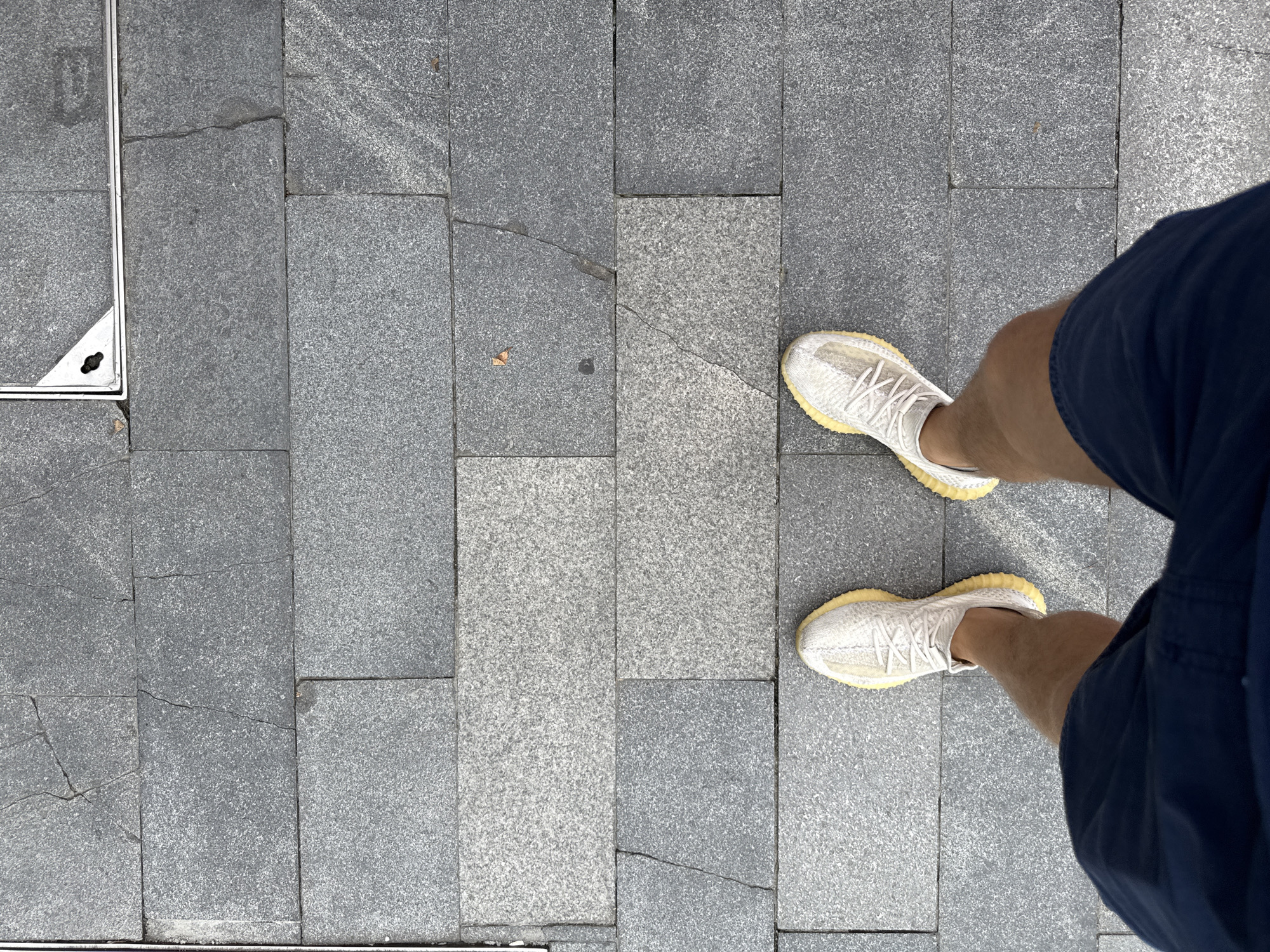The image shows a first-person perspective of a person standing on a sidewalk in Haizhu District, China. The sidewalk is paved with large, rectangular gray granite tiles. The tiles exhibit some cracks and variations in color and texture. In the upper portion of the image, a metal grate is visible, set into the pavement.
The person is wearing dark blue shorts and white athletic shoes with yellow soles. The shoes appear to be a popular style, possibly Adidas Yeezy Boost 350 V2. The person's legs and shorts are visible, suggesting they are standing still. The lighting indicates it is daytime, with no harsh shadows. The overall scene is mundane, depicting a common urban streetscape.