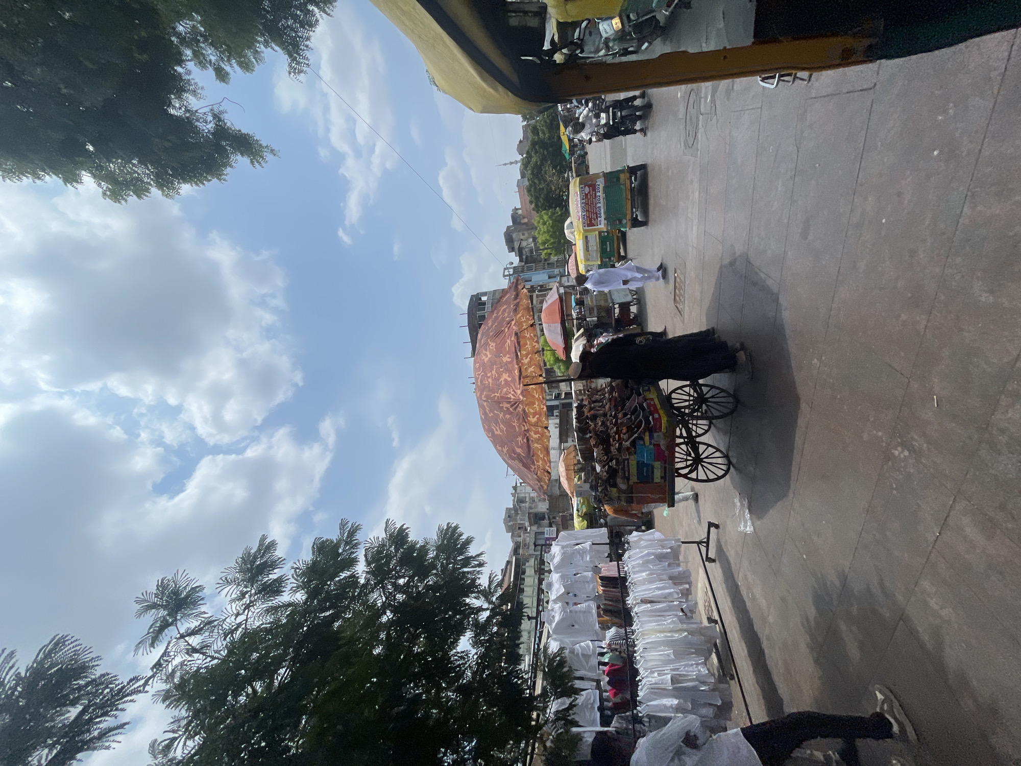 The image captures a street scene in Ahmedabad, India, under a bright, cloudy sky. In the foreground, a person in a white shirt and dark pants stands facing left, their arms crossed. To their right, a vendor pushes a cart filled with goods, sheltered by a large, patterned umbrella. The cart displays an assortment of items, possibly food or merchandise. Racks of white shirts hang to the left of the vendor, suggesting a clothing market. Further down the street, several auto-rickshaws are visible, some occupied by people, indicating bustling city life. Buildings with traditional Indian architecture line the background. The overall scene depicts a busy marketplace environment typical of Ahmedabad.