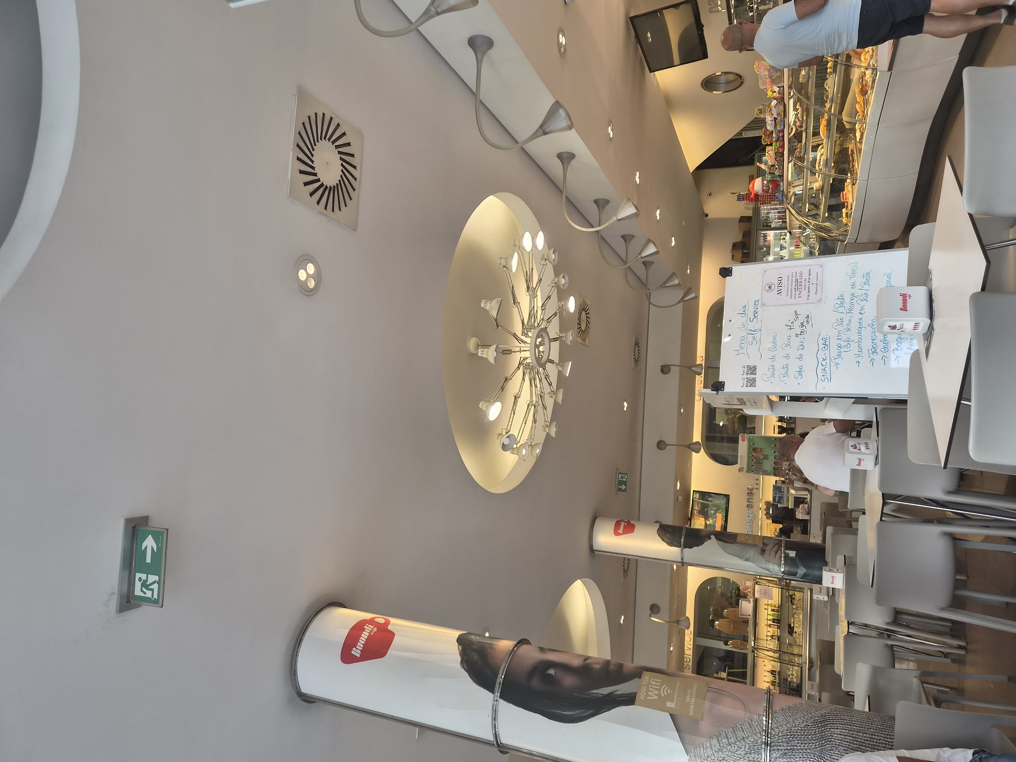 The image depicts the interior of a cafe or restaurant in Braga, Portugal. In the foreground, there are several tables and chairs, some of which are empty and some occupied by patrons. A whiteboard menu is visible on a table on the left. In the background, there is a display case filled with food items and a counter with staff serving customers. The cafe features modern lighting fixtures and decorative elements. An exit sign is mounted on the ceiling, and a ventilation grate is also visible. The overall ambiance appears to be casual and inviting.