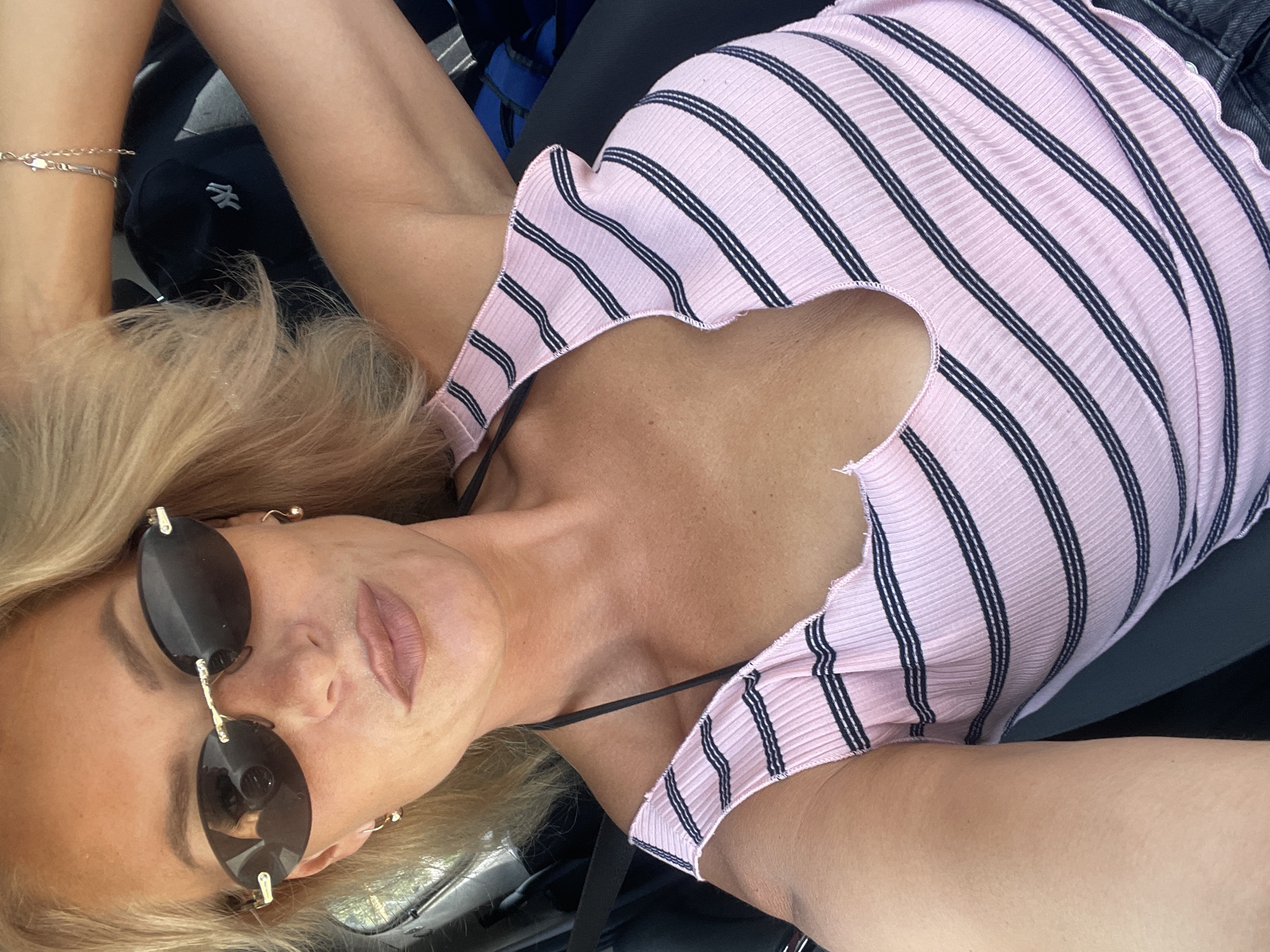 A blonde woman with fair skin and rosy cheeks is captured in a close-up, selfie-style shot. She is wearing sunglasses with dark, oval lenses and gold trim, and small gold earrings. Her lips are painted with a matte, nude lipstick. Her hair is styled in loose waves, framing her face.
She is wearing a pink and black striped ribbed tank top with a low neckline and thin black straps. The top appears to be made of a stretchy material, and the hemline is wavy. A black bra strap is visible under the tank top. Her right arm is raised, and a delicate silver bracelet with geometric charms is visible on her wrist.
The background suggests she is in a vehicle. A black baseball cap with a white logo (possibly the New York Yankees logo) is visible behind her. The seatbelt is fastened across her chest. The lighting indicates it is daytime, with natural light illuminating the scene. The environment does not provide any specific details about Baranovka, Russia.