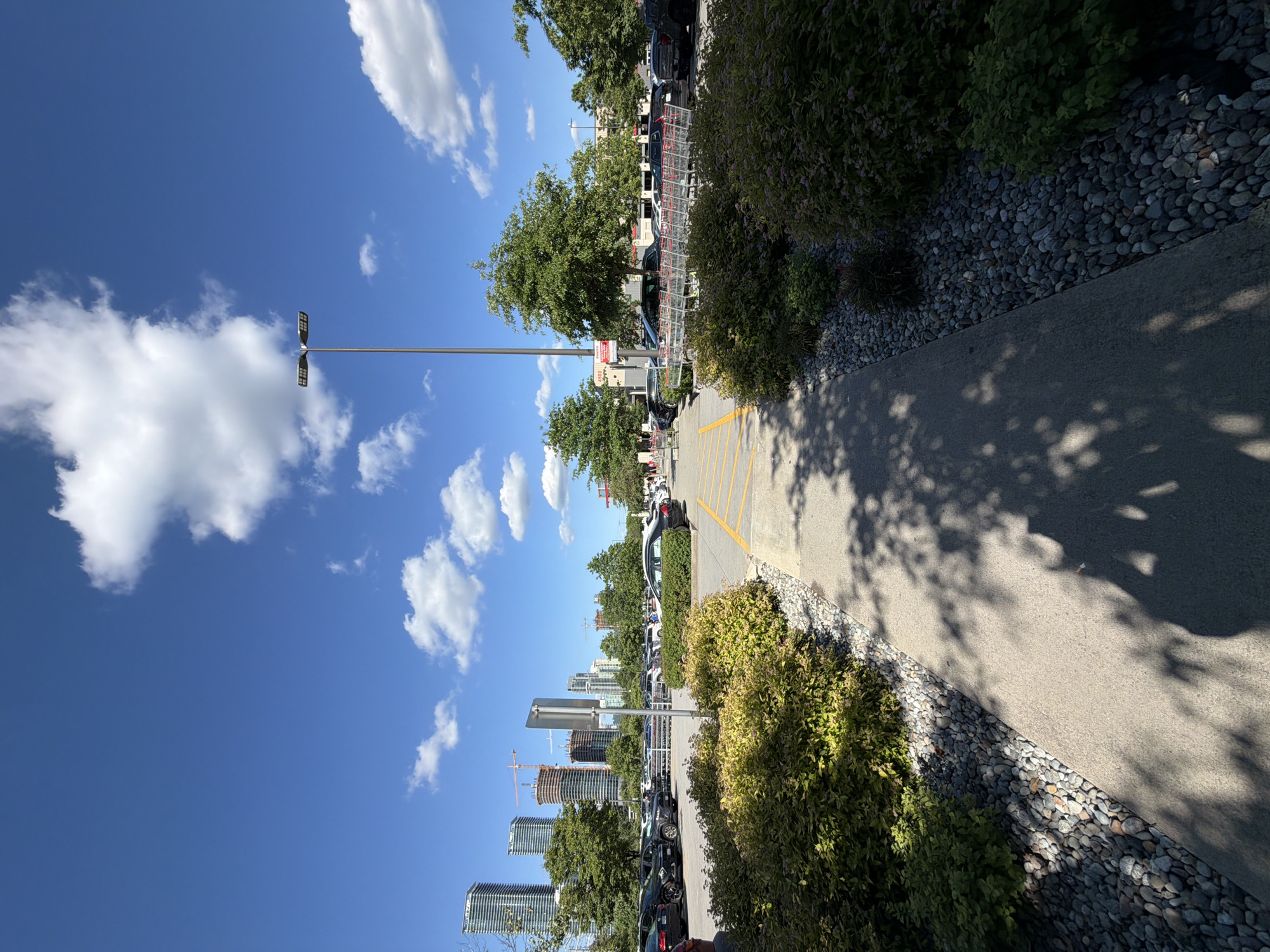 This is a wide-angle, eye-level shot of a shopping cart corral in a parking lot outside of a retail store. The scene is set in Burnaby, Canada, with a clear blue sky and scattered white clouds above. In the background, several tall, modern buildings and construction cranes are visible, indicating an urban environment.
In the foreground, a paved walkway is bordered by lush green bushes and grey gravel. The walkway leads towards the shopping cart corral, where numerous red shopping carts are neatly lined up. Several cars are parked in the surrounding parking lot, and some individuals can be seen walking in the distance. The overall impression is a sunny day in a busy commercial area.