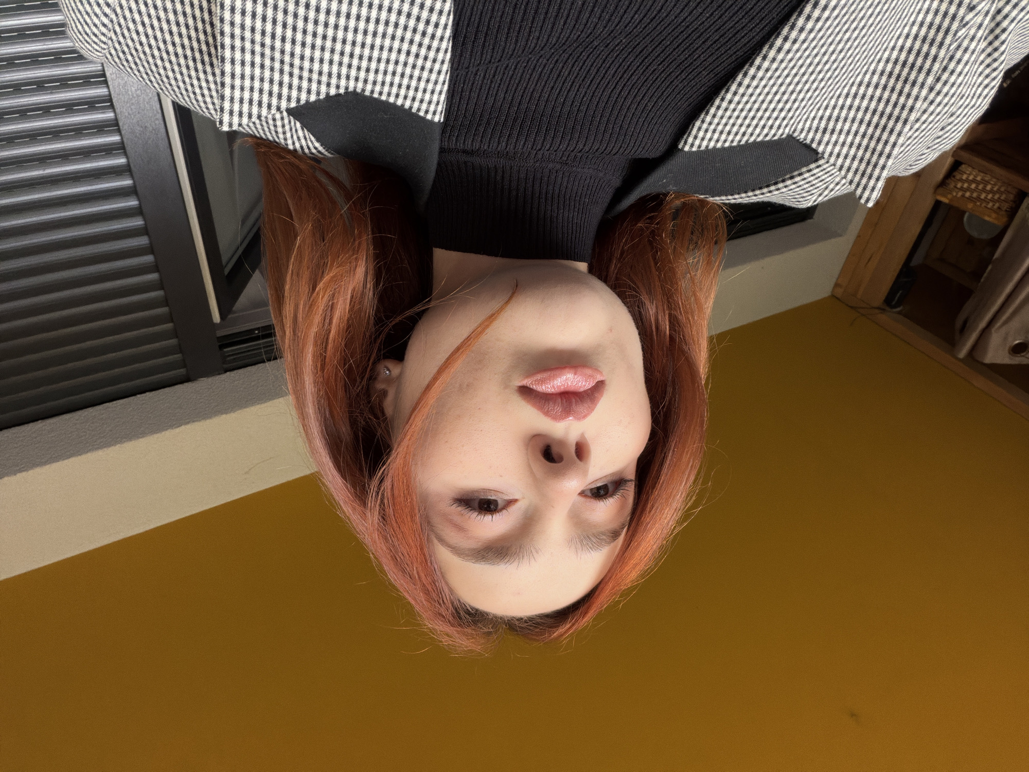 The image shows a person with auburn hair, wearing a black turtleneck sweater and a black and white checkered jacket, looking down at the camera. The background includes a window with closed blinds and a yellow wall. The location is not discernible from the image.