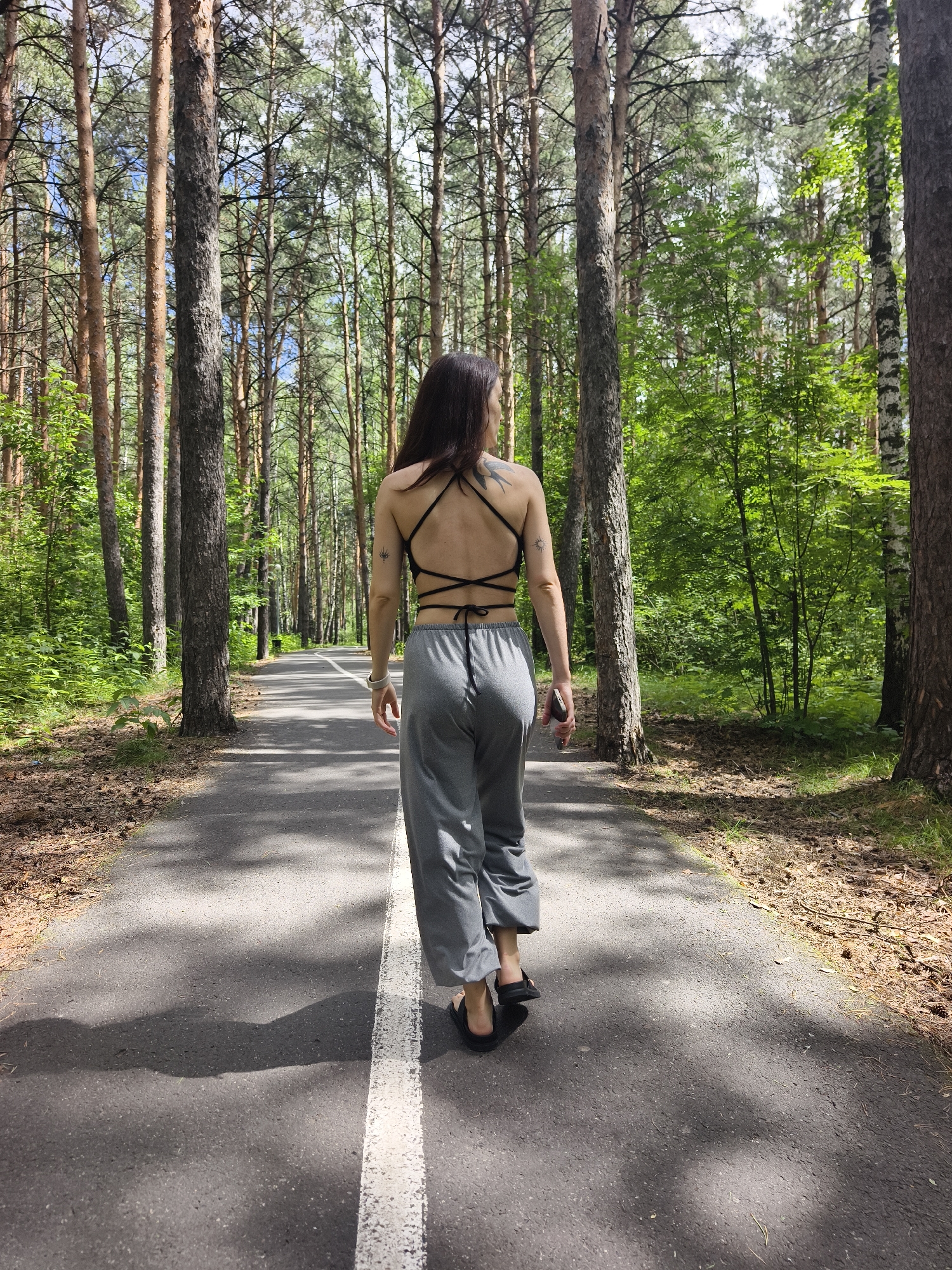 This media file features a rear view of a woman walking away from the camera on a paved path in a forest. The woman has long, dark hair and is wearing a black halter top and loose gray pants. She has tattoos on her upper back and arm. She is wearing black sandals and appears to be holding a dark object, possibly a phone, in her right hand.
The setting is a dense forest with tall pine trees and lush green undergrowth. The path is asphalt with a white line down the center. Sunlight filters through the trees, casting dappled shadows on the path. The overall mood is peaceful and serene. The scene suggests it is daytime, likely late morning or early afternoon, given the quality of the light. The location is identified as Tyumen, Russia, which is consistent with the type of forest depicted. There is no visible text in the image.