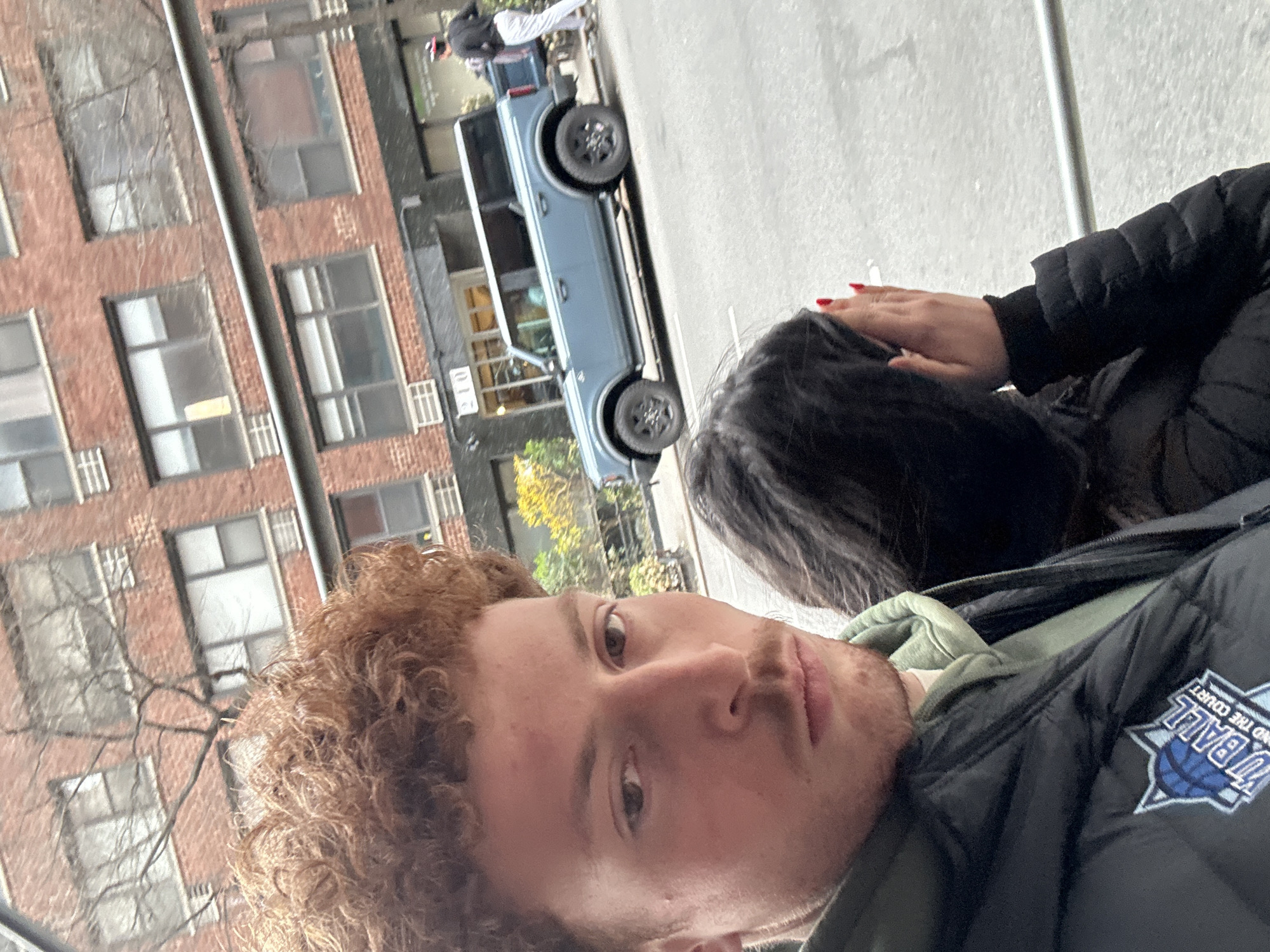 The image depicts an outdoor urban street scene in New York City, specifically identifiable by the "210" signage on a prominent brick building, strongly indicating the vicinity of 210 East 22nd Street in Manhattan's Gramercy Park neighborhood.
In the foreground, two individuals are present. On the left, an adult male with light skin, reddish-brown curly hair, and light stubble stares directly at the camera. He wears a black puffer vest bearing a blue and white "TU BALL AND THE COURT" logo over a light green hooded sweatshirt. To his right, a second individual, largely obscured, has dark hair. Their right hand, with red-painted fingernails, is raised to cover part of their face, appearing to hold a lit cigarette. This person is wearing a dark puffer jacket.
In the mid-ground, a light blue, four-door Ford Bronco SUV with a white roof and black wheels is parked parallel to the curb on a grey asphalt street. A silver horizontal metal bar extends across the frame in front of the vehicle.
The background features a multi-story red brick building with numerous windows and visible air conditioning units. The number "210" is clearly displayed above its ground-level entrance. A leafless tree stands in front of this building. Further down the street and to the right, another individual wearing a dark top, light pants, and a red and white baseball cap is walking away from the camera, seemingly interacting with the rear of the parked Bronco or carrying an item. The sky is overcast.
