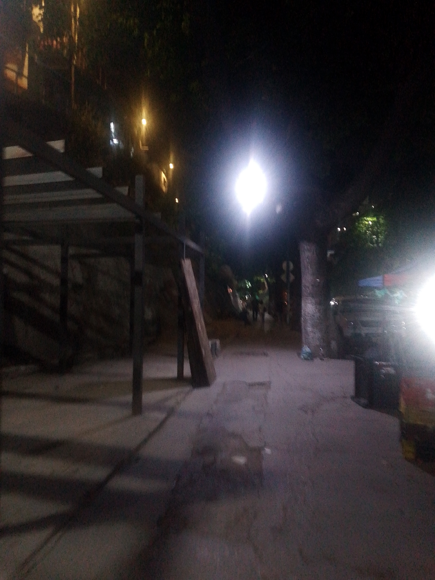 The image depicts a dimly lit street scene at night, possibly in a location in the city of Caracas, Venezuela. In the foreground, there is a dark metal structure, perhaps part of a bus stop or covered walkway, casting shadows on the ground. Further down the street, there is a bright street light. A few individuals appear to be walking in the distance, their forms indistinct due to the low light. A sign can be discerned near the right edge of the road. A vehicle or cart is visible to the right. The overall impression is of a quiet, poorly lit street.
