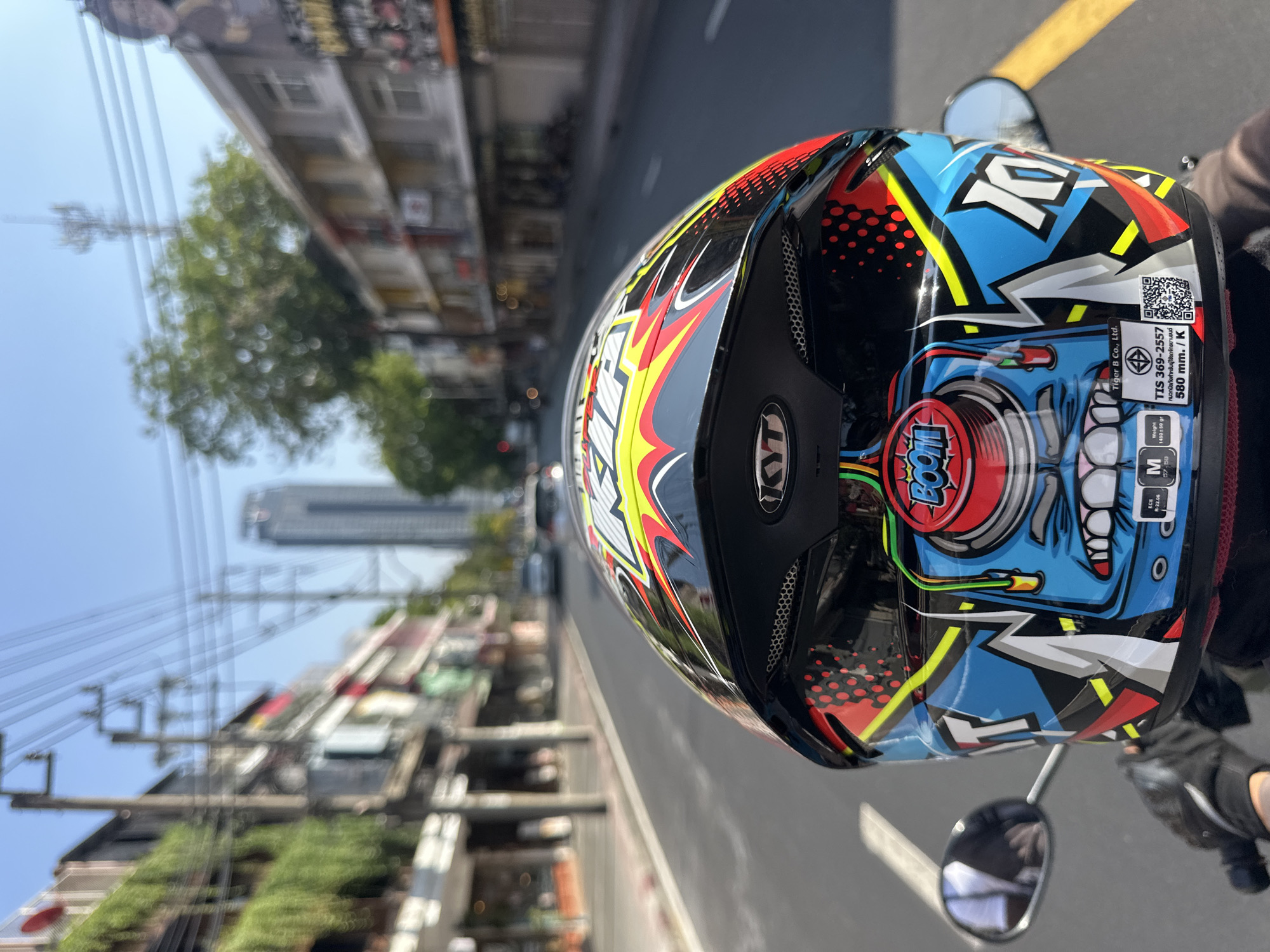 Here is a concise description of the image:
The image shows the back of a motorcycle helmet with a colorful design including the word "BOOM!" The helmet has a certification sticker and size information, indicating a medium size. The background is blurred, but it appears to be a street in an urban environment, likely Bangkok, Thailand, as indicated by the Thai script on the helmet's sticker. The buildings, trees, and power lines suggest a typical city street scene, with a taller building visible in the distance. A motorcycle mirror is also visible.