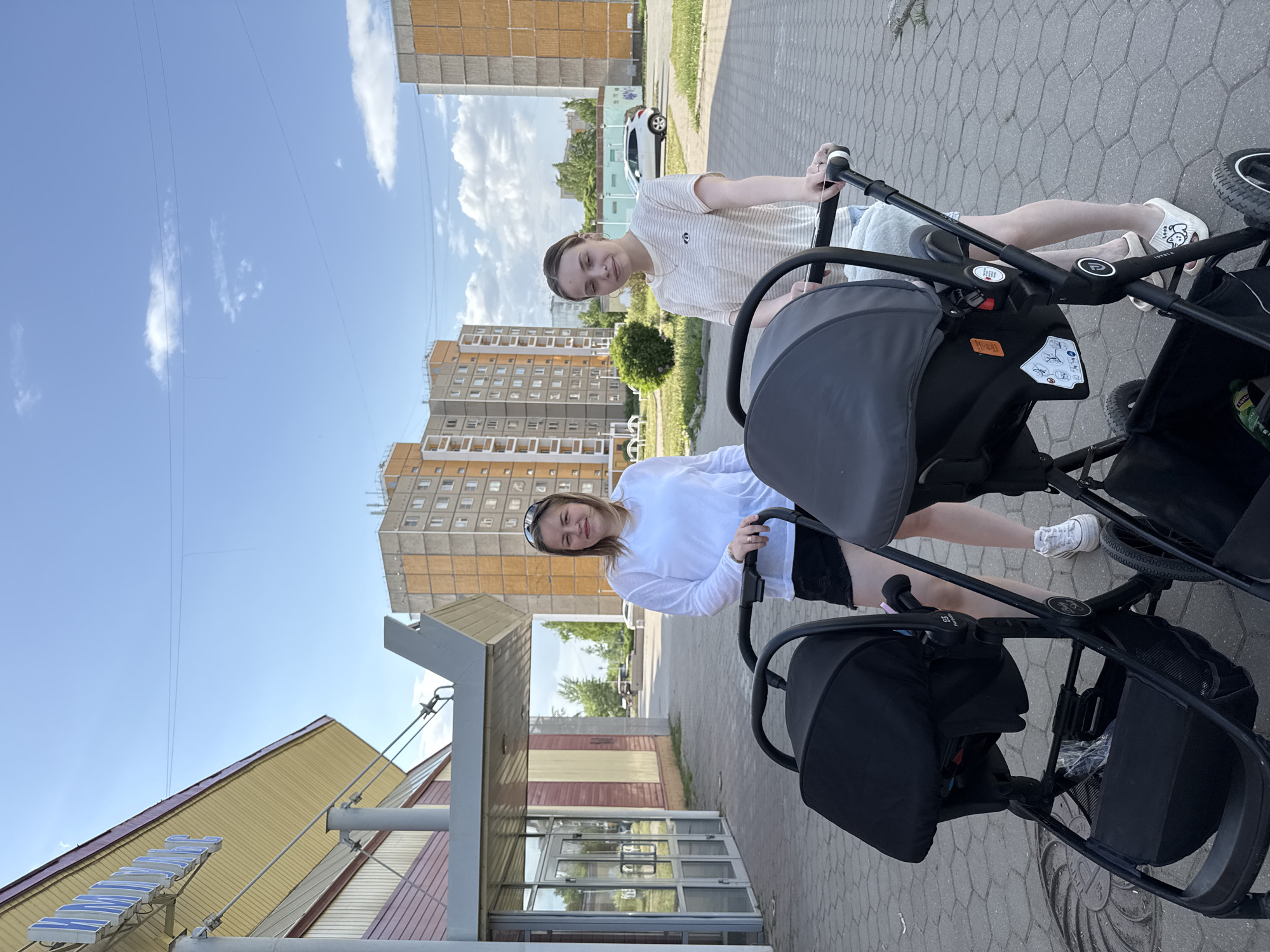 Two young women are standing outside a building with baby carriages in the city of Chaikovsky, Russia. The woman in the front is wearing a white long-sleeved shirt and black shorts. The woman behind her is wearing a white and tan striped shirt and light-colored shorts. Both are smiling. The building they are in front of has a large glass facade. Behind them are concrete apartment buildings. The sky is blue with some scattered clouds. The ground is paved with gray bricks.