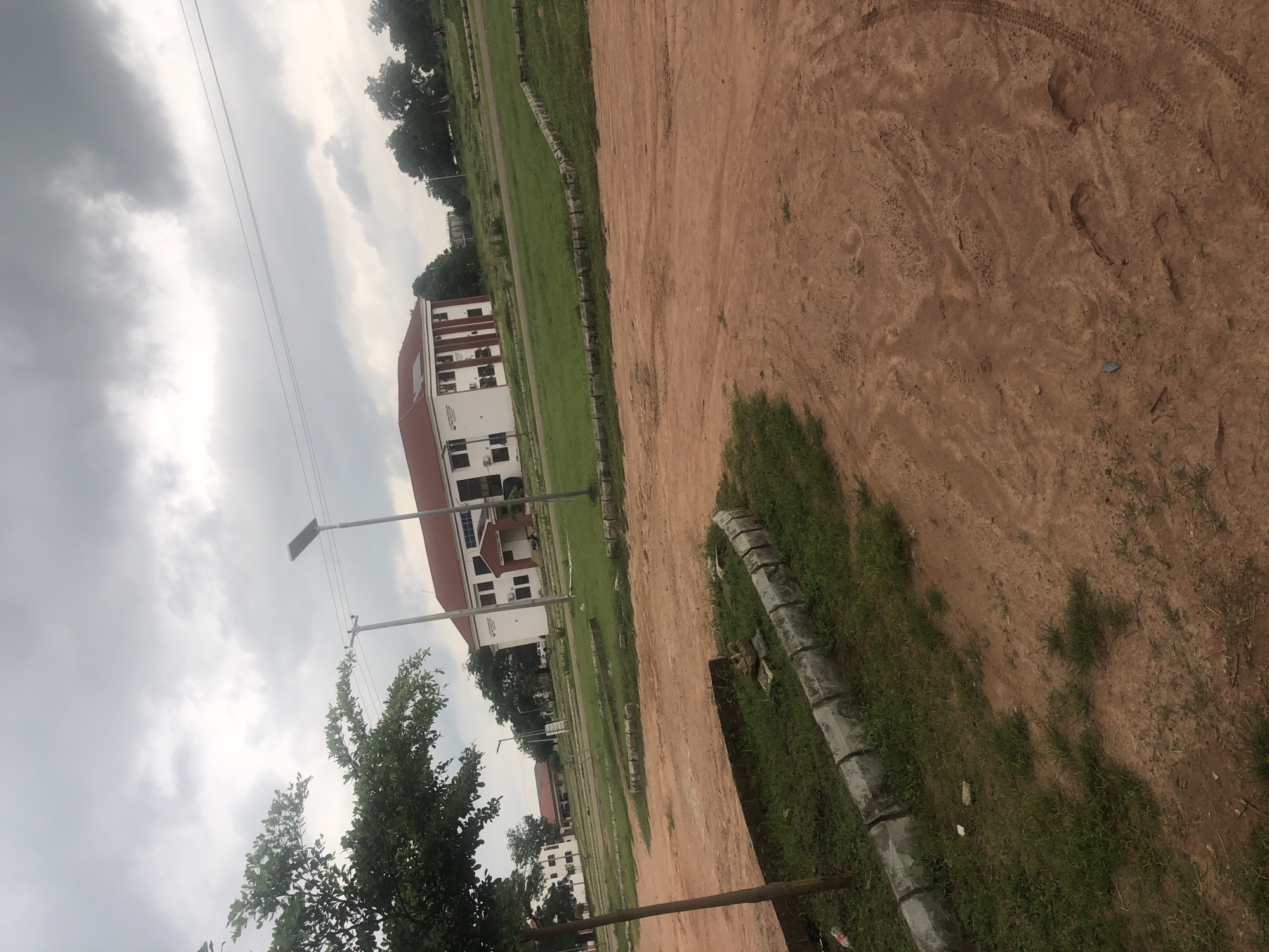 An outdoor scene in Wukari, Nigeria, depicts a modern, multi-story building with a red tiled roof and white walls, featuring large windows and vertical maroon accents. The building appears to be an institutional or educational facility, possibly a university complex, with a sign visible on its facade. In front of the building, a well-maintained lawn stretches out, bordered by a decorative stone wall. To the left of the frame, a tree with lush green foliage is prominently featured, its trunk a light brown color. The ground in the foreground is a reddish-brown dirt path, showing tire tracks and footprints, suggesting vehicular and foot traffic. Several lampposts, some with solar panels, are visible throughout the scene, indicating an emphasis on modern infrastructure and lighting. The sky is overcast with gray clouds, and the overall environment suggests a daytime scene in a developed area with greenery and contemporary architecture.
