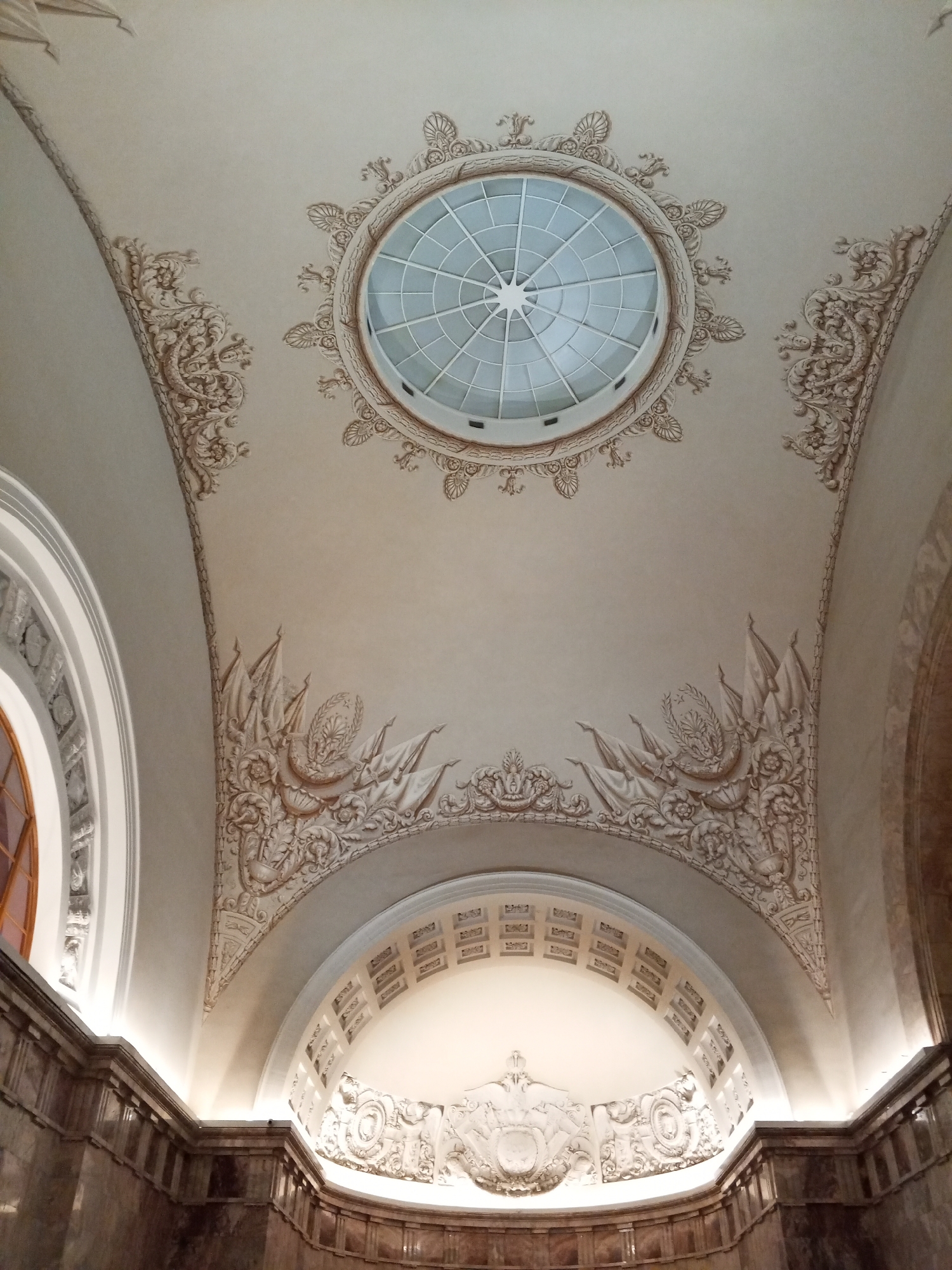 The image shows the interior of a building in Saint Petersburg, Russia, specifically the vestibule of the Kirovsky Zavod metro station. The scene focuses on the ceiling's architecture and ornamentation. The ceiling is arched and adorned with intricate bas-reliefs featuring floral patterns, flags, and other classical motifs. A central, circular skylight with a grid pattern is visible at the top of the arch. The walls are paneled with dark marble, and the lower part of the arched wall displays more bas-reliefs. Artificial lighting illuminates the scene, highlighting the details of the ceiling and walls.