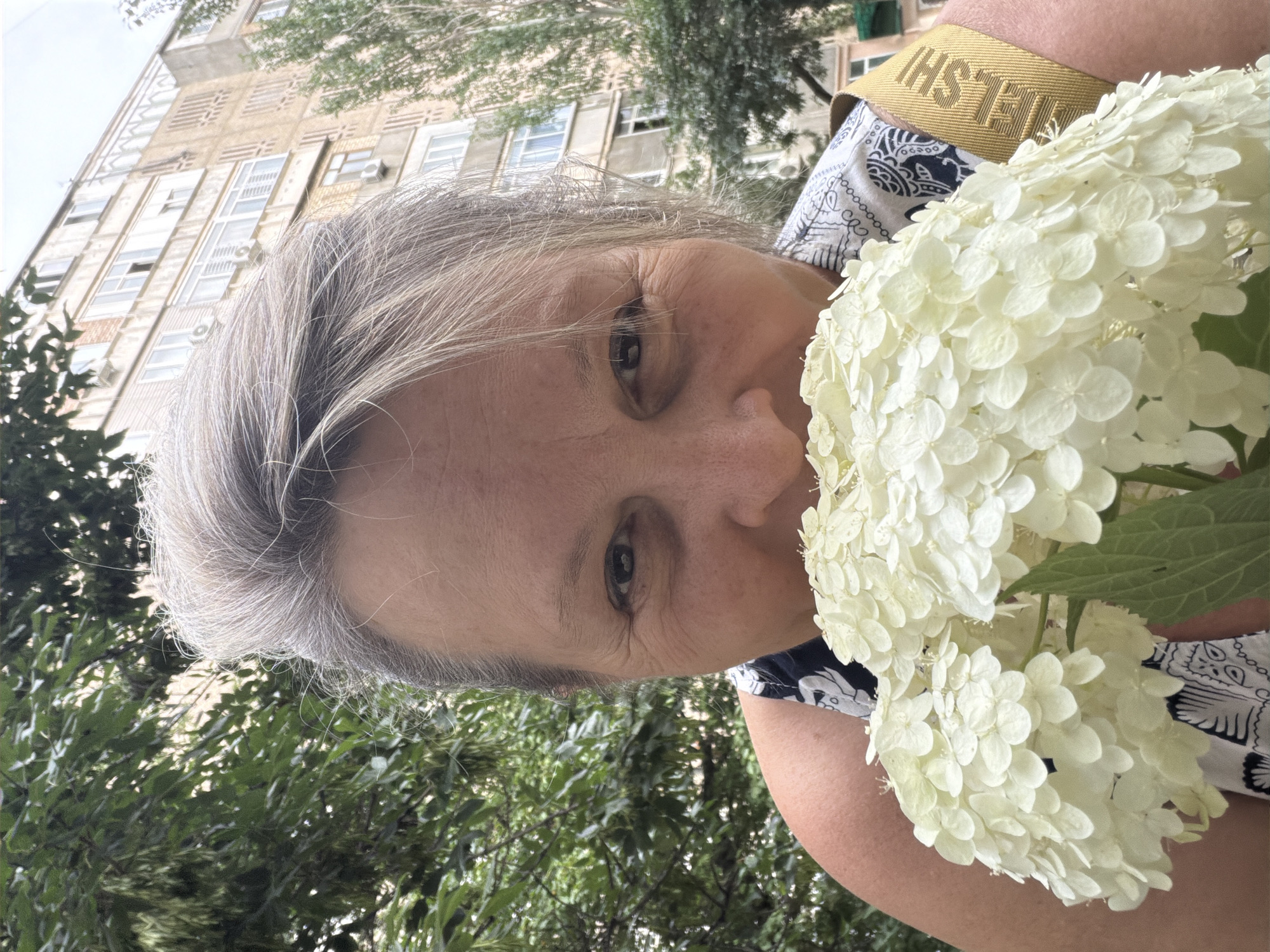 An over-the-shoulder, close-up, outdoor selfie shows a woman with graying hair holding a large cluster of white flowers to her face. She is wearing a dark patterned top and a yellow strap across her shoulder. In the background, a multi-story building with numerous windows and air conditioning units is visible, along with green trees. The setting appears to be an urban residential area.