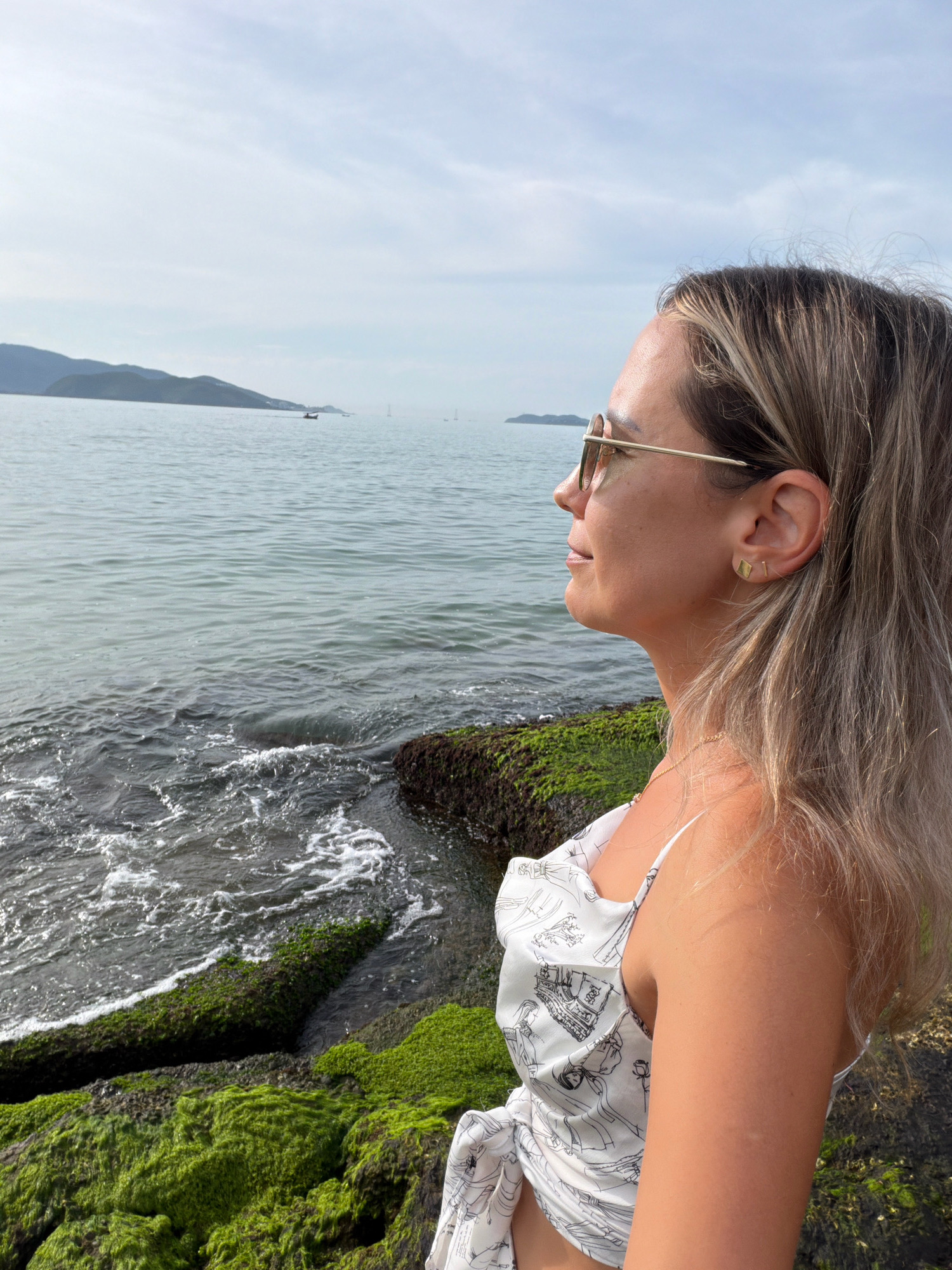 The image captures a woman in the foreground, facing to the right, with the ocean and a hazy coastline in the background. The woman has blonde hair, is wearing sunglasses and a white top with a black line drawing pattern. She is standing on rocks covered in green algae. The ocean is calm with gentle waves lapping at the shore. In the distance, hills and mountains are visible, and there are a few small boats on the water. The sky is clear with some wispy clouds. This scene is likely located along the coast of Nha Trang, Vietnam, known for its beaches and mountainous backdrop.