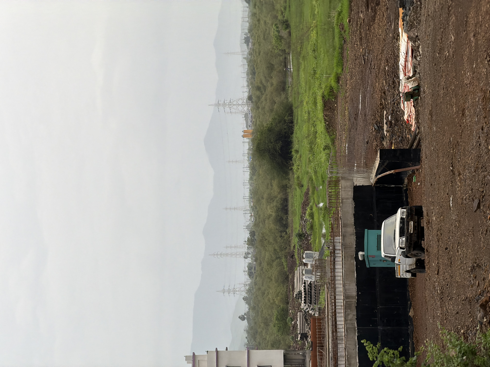 The image depicts a construction site in Vasai-Virar, India, on a cloudy day. In the foreground, a dirt area shows a white pickup truck with a green generator behind it. A partially constructed bridge or elevated structure runs across the middle ground. Beyond the bridge, a dense area of green vegetation and trees extends towards the horizon. In the background, a line of high-voltage electricity pylons are visible against a hazy mountain range under an overcast sky. The overall impression is one of ongoing development in a semi-rural or suburban setting.