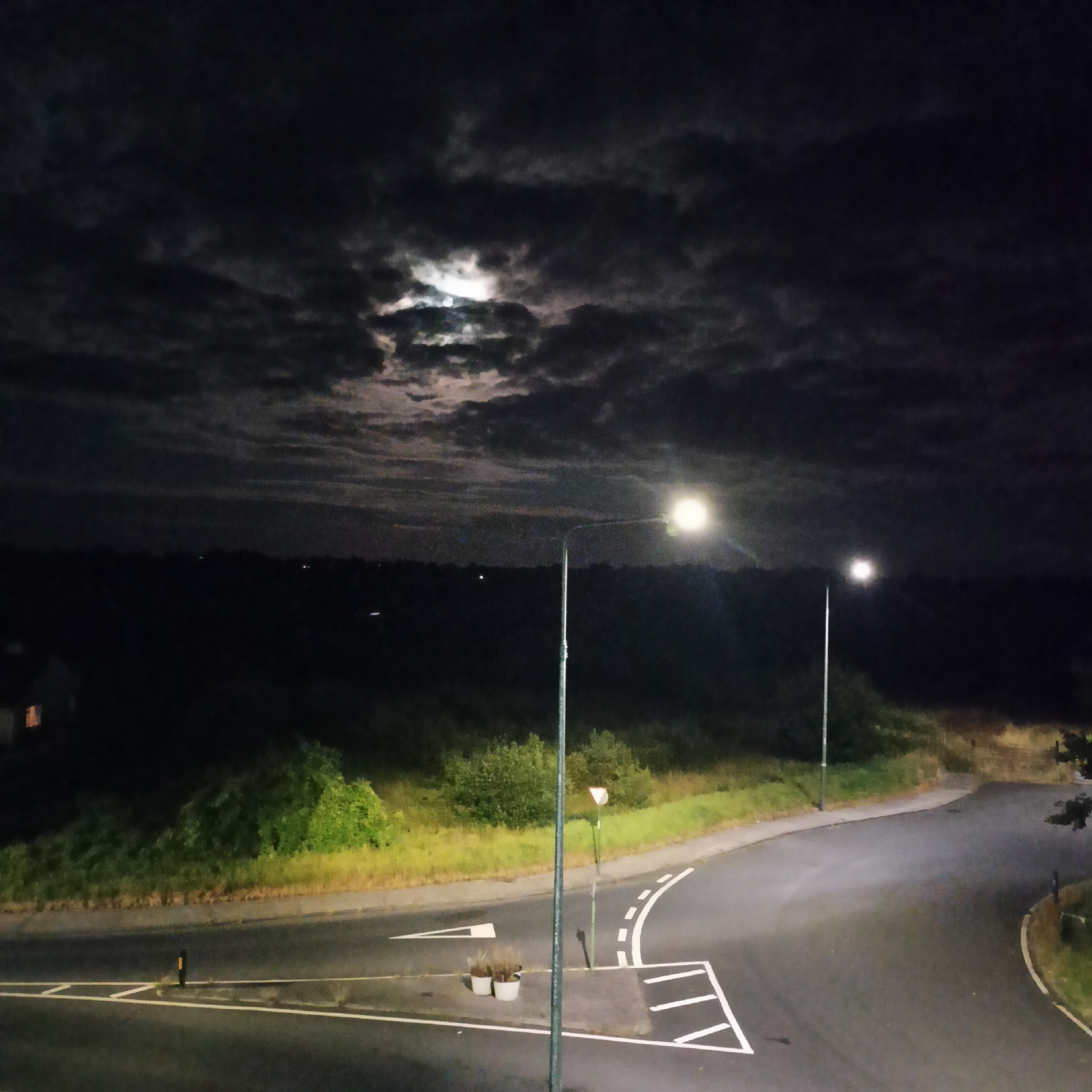 The image shows a nighttime scene in a rural or semi-rural area, possibly on the outskirts of Limerick, Ireland. A crescent moon is visible through scattered clouds in the dark sky. Two streetlights illuminate a paved road that curves to the right. The road has white lane markings, including directional arrows and a hatched area. There is vegetation on the left and right sides of the road, with a small building visible in the far left background. Two potted plants sit on a sidewalk area near one of the streetlights. The overall impression is one of a quiet, dimly lit landscape.