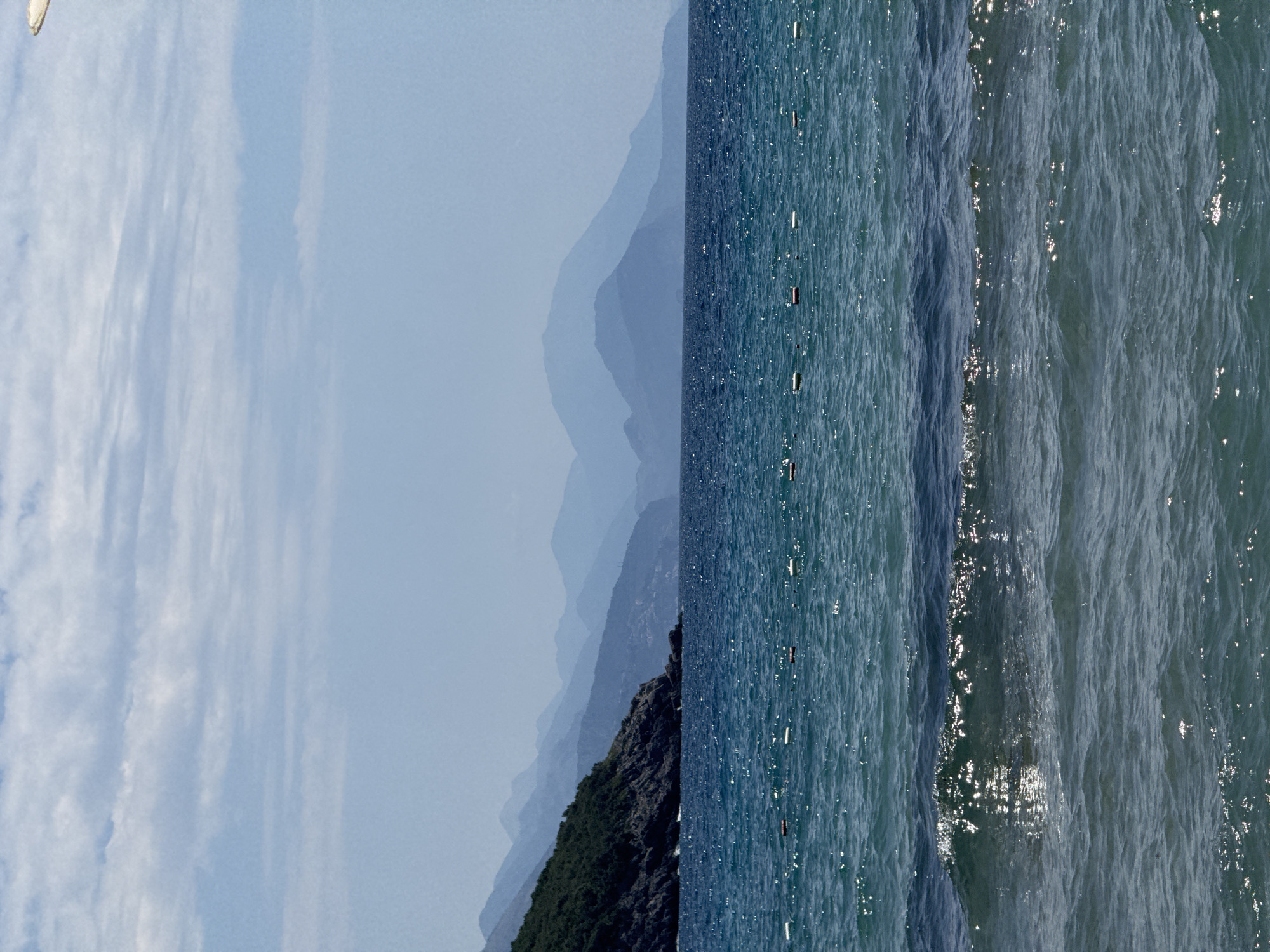 This is a landscape photograph of a coastal scene under a blue sky with scattered clouds. In the foreground, the ocean displays various shades of blue and green, with small waves reflecting sunlight. A line of buoys is visible across the water. In the middle ground, a rocky, tree-covered hillside juts out into the sea on the left. In the background, a series of progressively lighter blue mountains recede into the distance. The scene is devoid of people and any discernable man-made structures other than the buoys.