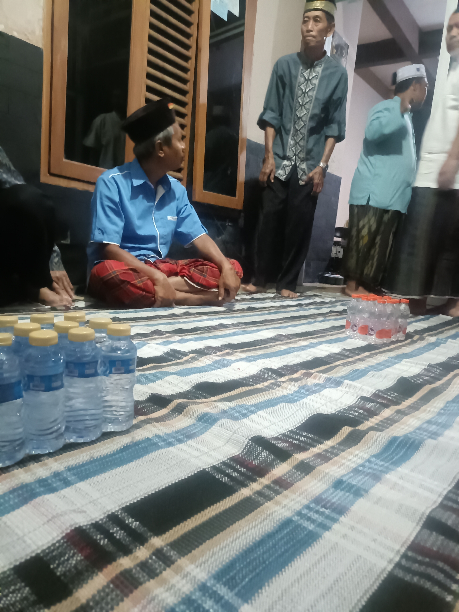 The image depicts an indoor scene where several individuals are gathered. A man wearing a blue shirt and a black peci hat is seated on a patterned mat, his legs crossed. To his right, another man in a teal shirt with intricate patterns and a golden-rimmed peci stands, looking to his left. Behind him, a third man, partially visible, wears a light blue shirt and a white peci, also with a patterned sarong. In the foreground, multiple bottles of water are arranged on the mat. The background shows wooden window frames and a dark wall. The scene is likely a gathering in a home or community space in Kediri City, Indonesia, possibly for a religious or social event.