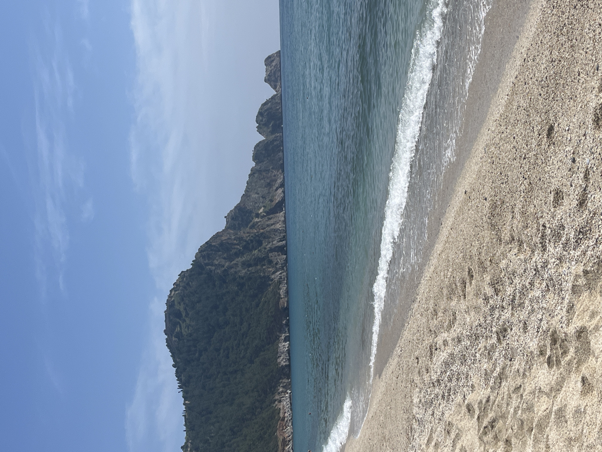 The image displays a serene coastal landscape oriented horizontally. On the left, a large, steep mountain or cliff face dominates the background, covered in dark green vegetation, indicating dense forestation, with some rocky outcrops visible. The sky above is a clear, bright blue with faint, wispy white clouds.
In the midground, the calm, blue-green waters of the sea stretch across, meeting the base of the mountain and extending towards the foreground. Gentle waves break along the shore, creating lines of white foam. A few very small, indistinct dark figures are visible in the water, suggesting people are swimming or wading.
The right side and foreground of the image feature a wide expanse of beach composed of light tan sand mixed with small pebbles. Footprints are discernible on the sand, indicating recent activity. The bright light suggests it is daytime, likely morning or early afternoon, under sunny weather conditions. The scene evokes a sense of natural beauty and tranquility, characteristic of a coastal destination like Alanya, Turkey. No discernible text is visible in the image.