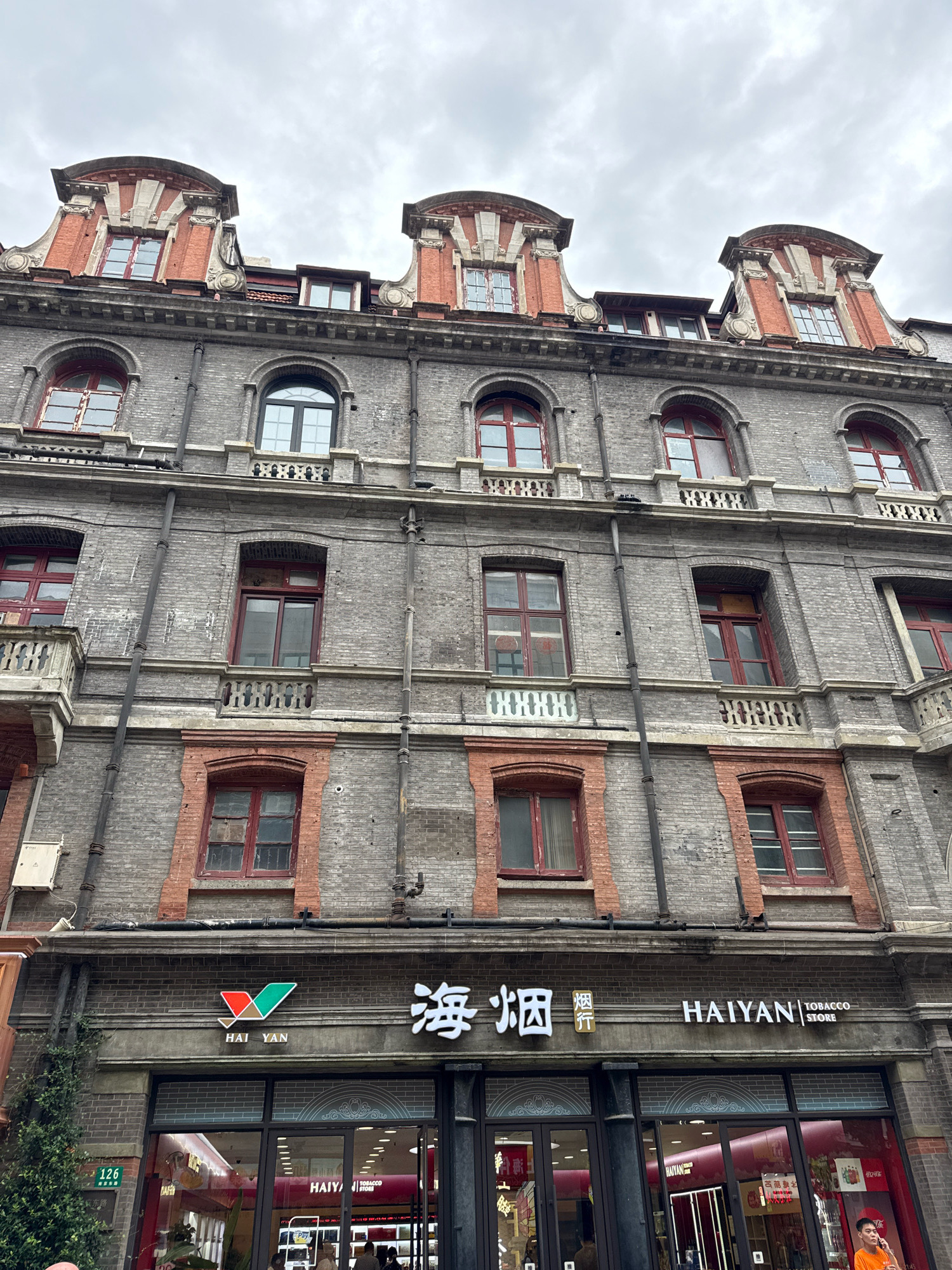 The image shows a low-angle, full-frame exterior shot of a multi-story brick building, likely in Shanghai, China, given the presence of Chinese characters on signage. The building features a mix of architectural styles, with ornate decorative elements on the roofline, including red brick gables with carved stone accents. The facade is constructed of grey brick, with numerous red-framed windows of various shapes and sizes distributed across several floors. Some windows are arched, others rectangular. Small, decorative balconies with balustrades are present on some levels. The ground floor of the building appears to be a commercial establishment, with large glass windows and signage that includes the words "HAIYAN TOBACCO STORE" and Chinese characters. A partial view of a street is visible at the bottom left with a green plant and a street number sign "126". The sky is overcast with clouds.