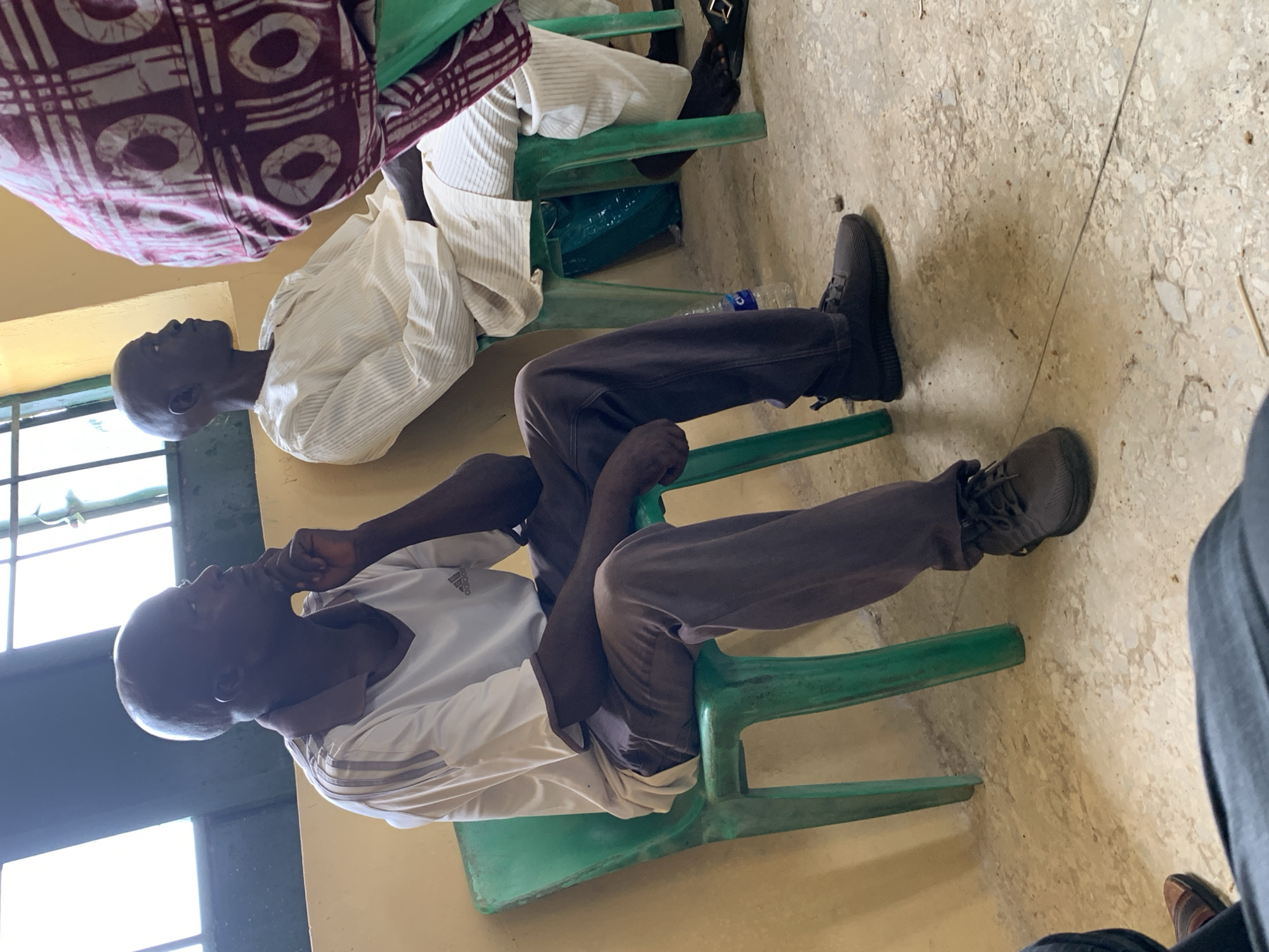 In a room in Akwanga, Nigeria, two adult men sit on light green plastic chairs. One man wears a white long-sleeved shirt and brown pants, talking on his phone. He also wears gray shoes. Another man, wearing a striped white robe, sits on another chair, seemingly listening. A patterned brown and white cloth is seen on the first chair, behind the second man. A window is partially visible in the background. The floor appears to be tiled.