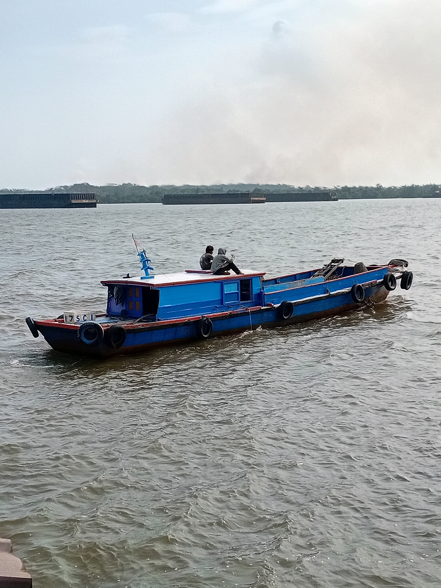 A blue and red boat with two individuals seated on its deck is in motion on a body of water, likely a river or strait, near Sungai Meriam, Indonesia. The boat has a cabin structure with a blue roof and a white upper section. The number "17SC.1" is visible on the bow of the boat. Several black tires are mounted along the sides and at the stern of the boat, likely as fenders. The water is choppy with visible waves. In the background, two large barges are stationary on the water, and a hazy, treed shoreline is visible beyond them. The sky is overcast with a light haze or smoke in the distance.