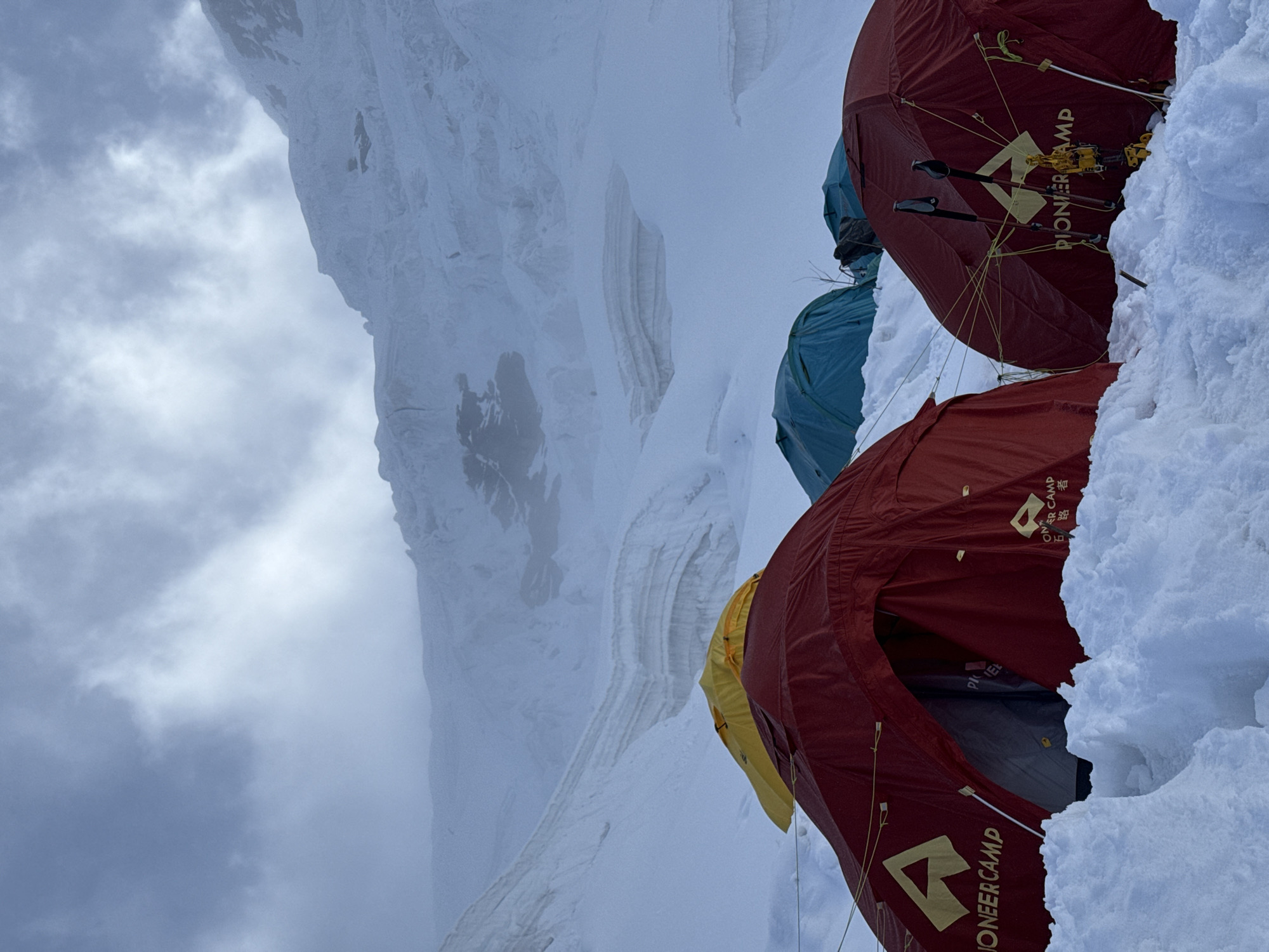 A high-altitude mountaineering camp is set up in the snow-covered mountains near Samagaun, Nepal. Several tents are pitched in the snow, including two prominent red tents in the foreground with "Pioneer Camp" branding. A yellow tent is visible behind the red tents. In the background, a large, snow-covered mountain range with steep ice walls and crevasses dominates the scene under a cloudy sky. Trekking poles are visible near the red tents.