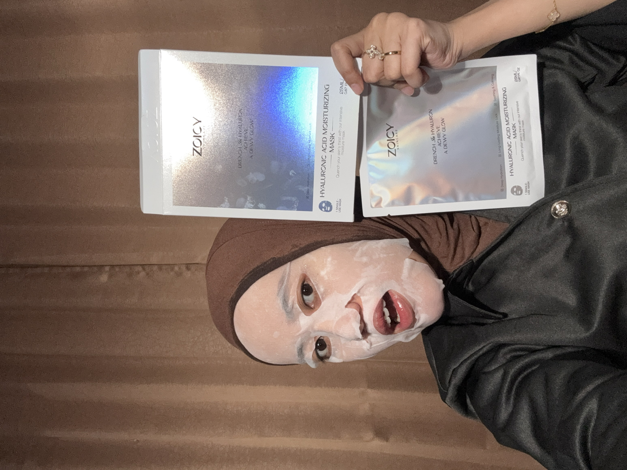 An indoor, sideways-oriented photograph shows a person wearing a white sheet mask on their face and a brown head covering. Their eyes are visible, looking towards the camera, and their mouth is slightly agape. They are wearing a dark, possibly satin or silky, long-sleeved garment. A gold-colored ring with a floral design is on a finger of their right hand, which holds a silver-holographic packet. A gold-colored bracelet with a clover charm is visible on their wrist.
The person holds two skincare products: a rectangular box and a single packet, both from the brand "ZOICY SKINCARE". The box, held upright by the person's left hand, is silver-holographic with text that includes "HYALURONIC ACID MOISTURIZING MASK," "DRENCH IN HYALURON ACHIEVE A DEWY GLOW," and "Deep Hydration." The packet, held by their right hand, shares a similar design and also states "HYALURONIC ACID MOISTURIZING MASK," "DRENCH IN HYALURON ACHIEVE A DEWY GLOW," "25ML / 0.87 FL OZ," "Deep Hydration," "Long Lasting Moisture Lock," "Soothing & Calming," and "1 SINGLE-USE MASK." The background features a textured brown surface, possibly a wall or curtain.
