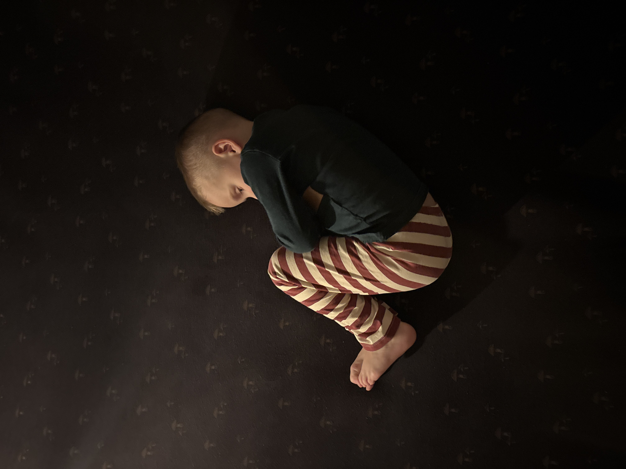 An overhead, dimly lit photograph depicts a young child, appearing to be a boy with short blonde hair, positioned on their left side in a curled, semi-fetal posture. The child wears a dark teal long-sleeved shirt and red and off-white striped trousers, with bare feet. Their arms are crossed over their chest, and their face is turned inwards, partially obscured. The child rests on a dark, possibly black, surface textured with a repeating pattern of small, faint, light-colored stylized mushroom shapes. Illumination originates from the upper left, casting a distinct shadow across the right side of the child and the background. The scene is an indoor domestic setting located within the city of Chaikovsky, Russia.