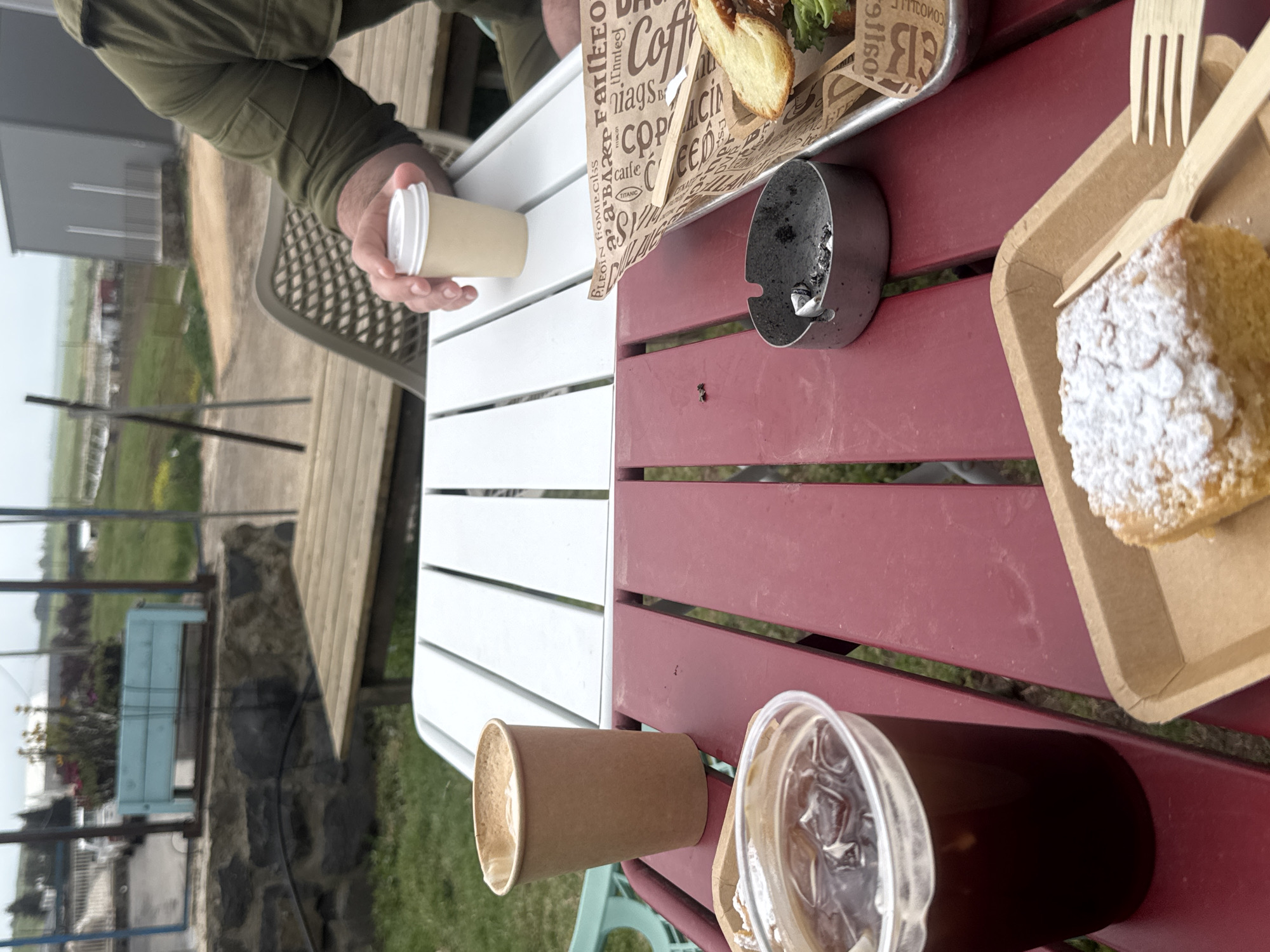 The image shows a table with food and drinks on it. The table is located outdoors, with a view of fields and trees. A person is holding a coffee cup. Other items on the table include a sandwich, cake, and a glass of coffee. There is an ashtray on the table with cigarette butts in it. The table is half white and half maroon. The image may have been taken in the Golan Regional Council, Israel, since the setting looks similar to its environment.