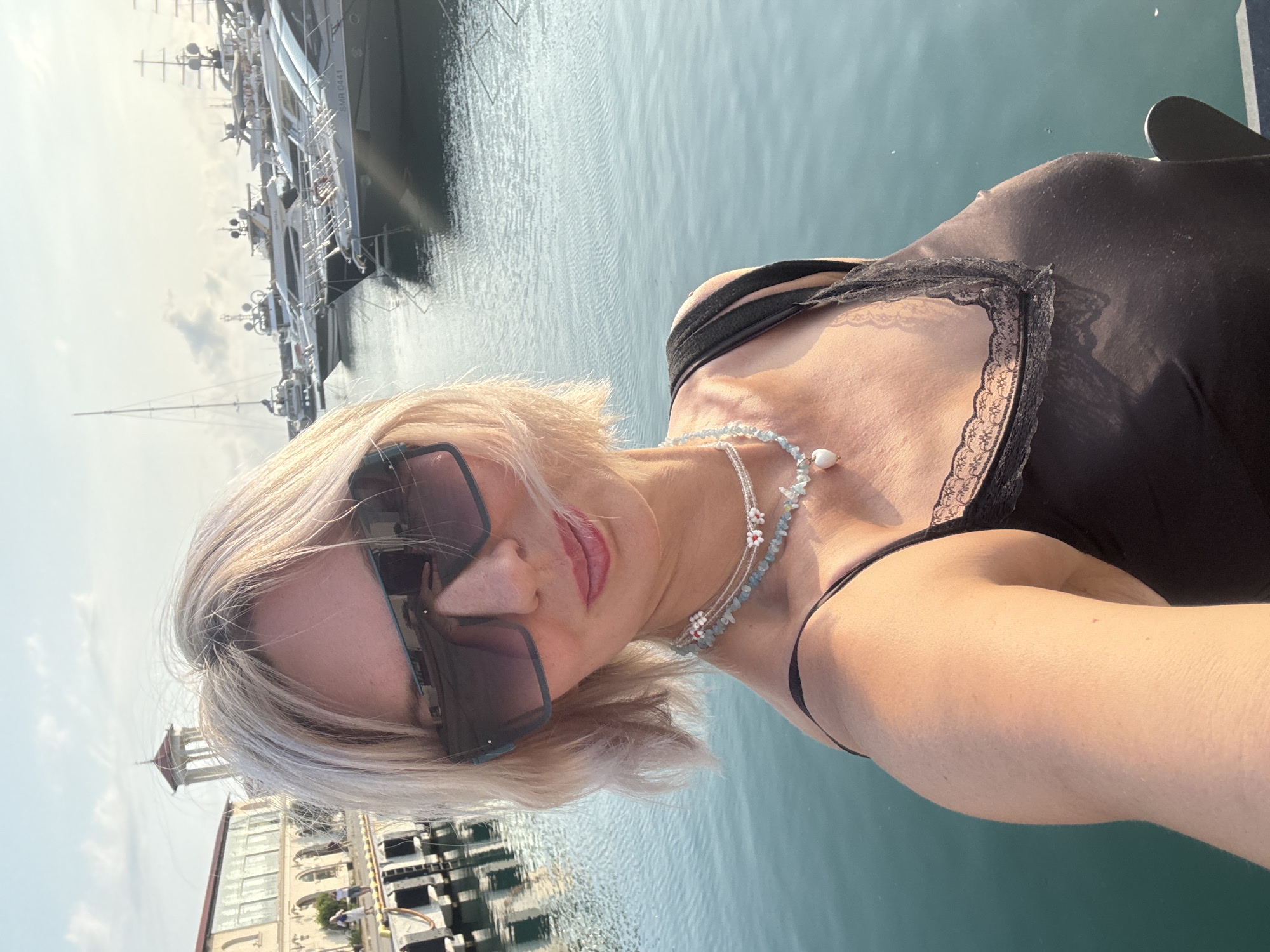 A woman with short, blonde hair is taking a selfie at a marina in Sochi, Russia. She is wearing large, dark sunglasses, a black tank top with lace trim, and a multi-layered beaded necklace. Behind her, several yachts are docked in the turquoise water. In the background, a building with arched windows and a tower can be seen, along with a clear sky.