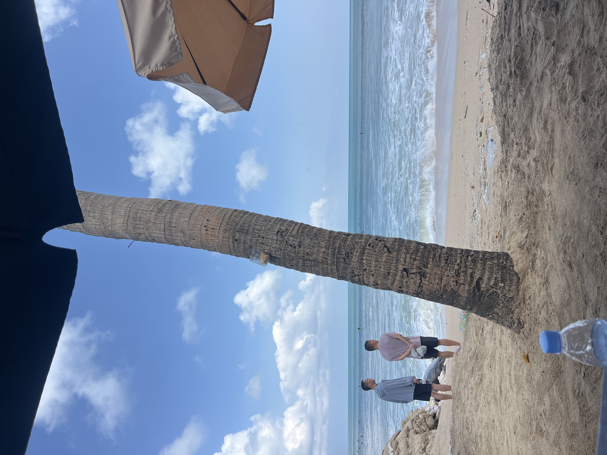 This is a wide-angle, eye-level shot of a beach in Kuta, Indonesia. The foreground is dominated by dark sand, with a clear plastic water bottle in focus. In the mid-ground, a sandy beach transitions to the ocean. Two men are standing on the beach, facing the ocean. The man on the left wears a grey hooded jacket and black shorts. The man on the right wears a light pink t-shirt and black shorts. Both men are holding clear plastic bags. A large palm tree trunk is prominent in the right mid-ground, with a plastic bag containing snacks attached to it. Beyond the palm tree, a light-colored beach umbrella is partially visible. The ocean is a bright turquoise, with white waves breaking on the shore. The sky is a vibrant blue with scattered white clouds. In the far distance, several people can be seen in the water, some appearing to be surfing. The overall scene suggests a typical beach day in Kuta.