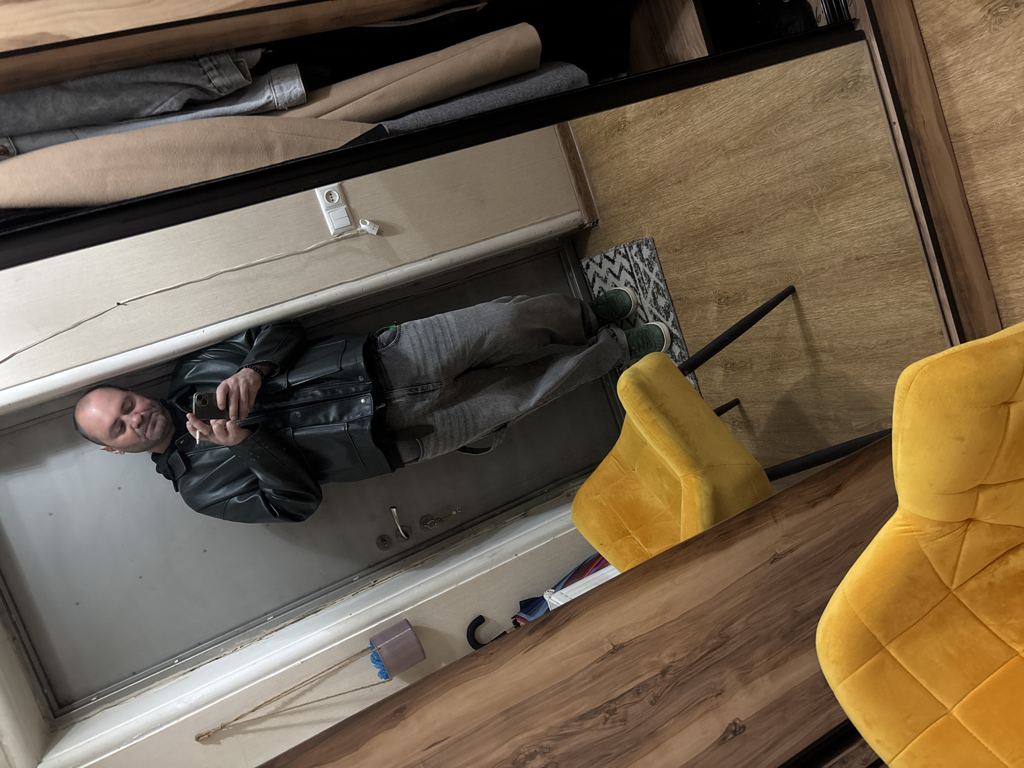 A man in a black leather jacket and gray pants stands in front of a closet mirror, holding a phone and a cigarette. He is reflected in the closet mirror. He is wearing green shoes. The closet is filled with clothes and miscellaneous items. A yellow chair sits nearby. The location appears to be indoors, in Tbilisi, Georgia.
