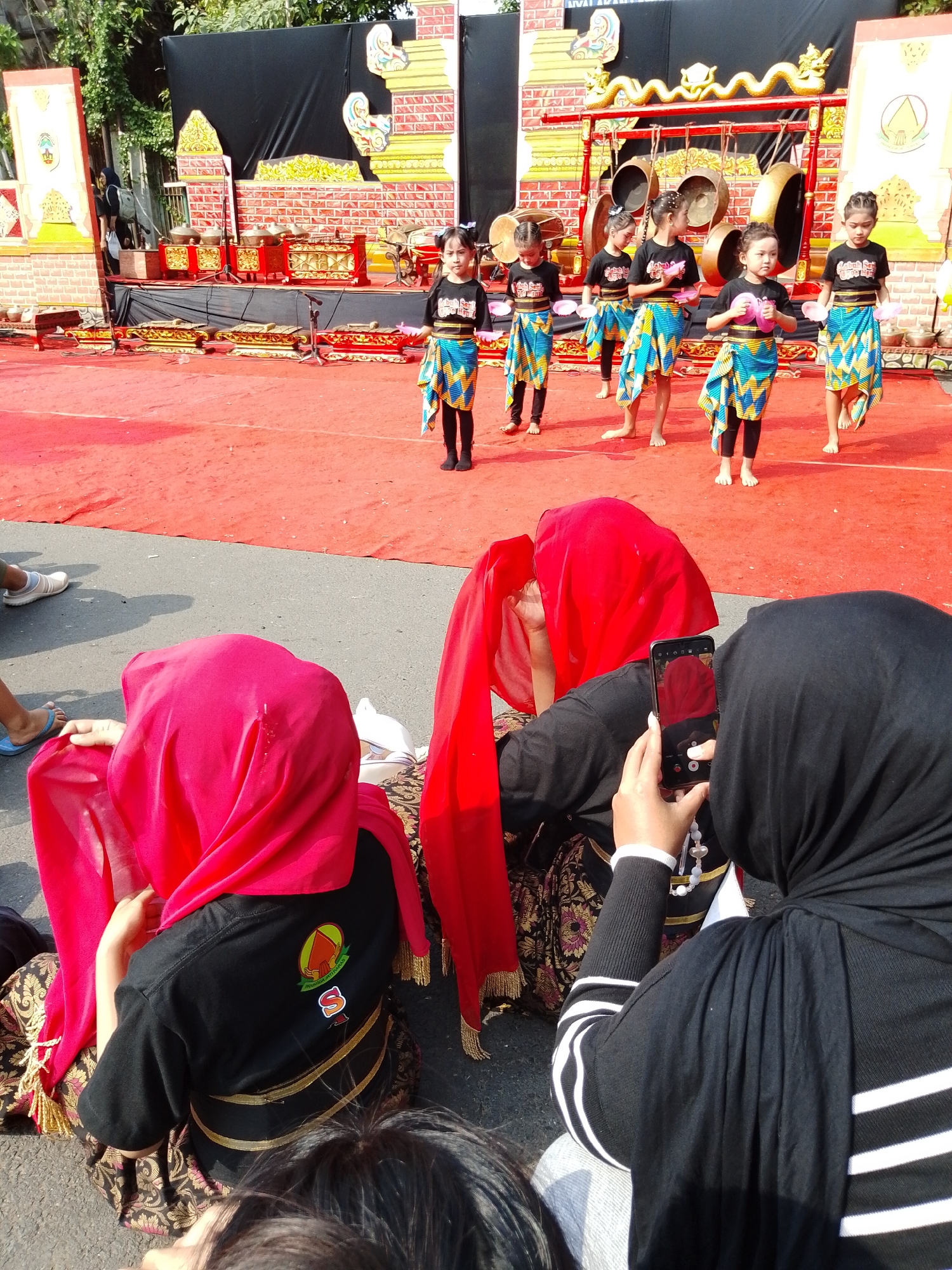 In a street scene in Kecamatan Pati, Indonesia, an outdoor performance is underway on a red carpeted stage. Several young girls, dressed in black tops and vibrant patterned skirts, stand on the stage. They hold pink objects, possibly flowers, in their hands. Behind them, a traditional Indonesian gamelan instrument setup, featuring large bronze gongs and wooden xylophones, is visible. The stage is decorated with colorful backdrops. In the foreground, two individuals are seated on the ground, facing away from the camera. One person is covered by a red headscarf and the other by a black hijab. The person in the black hijab is holding a smartphone and appears to be taking a photograph or recording a video of the performance. The overall environment suggests a cultural event taking place in a public area.