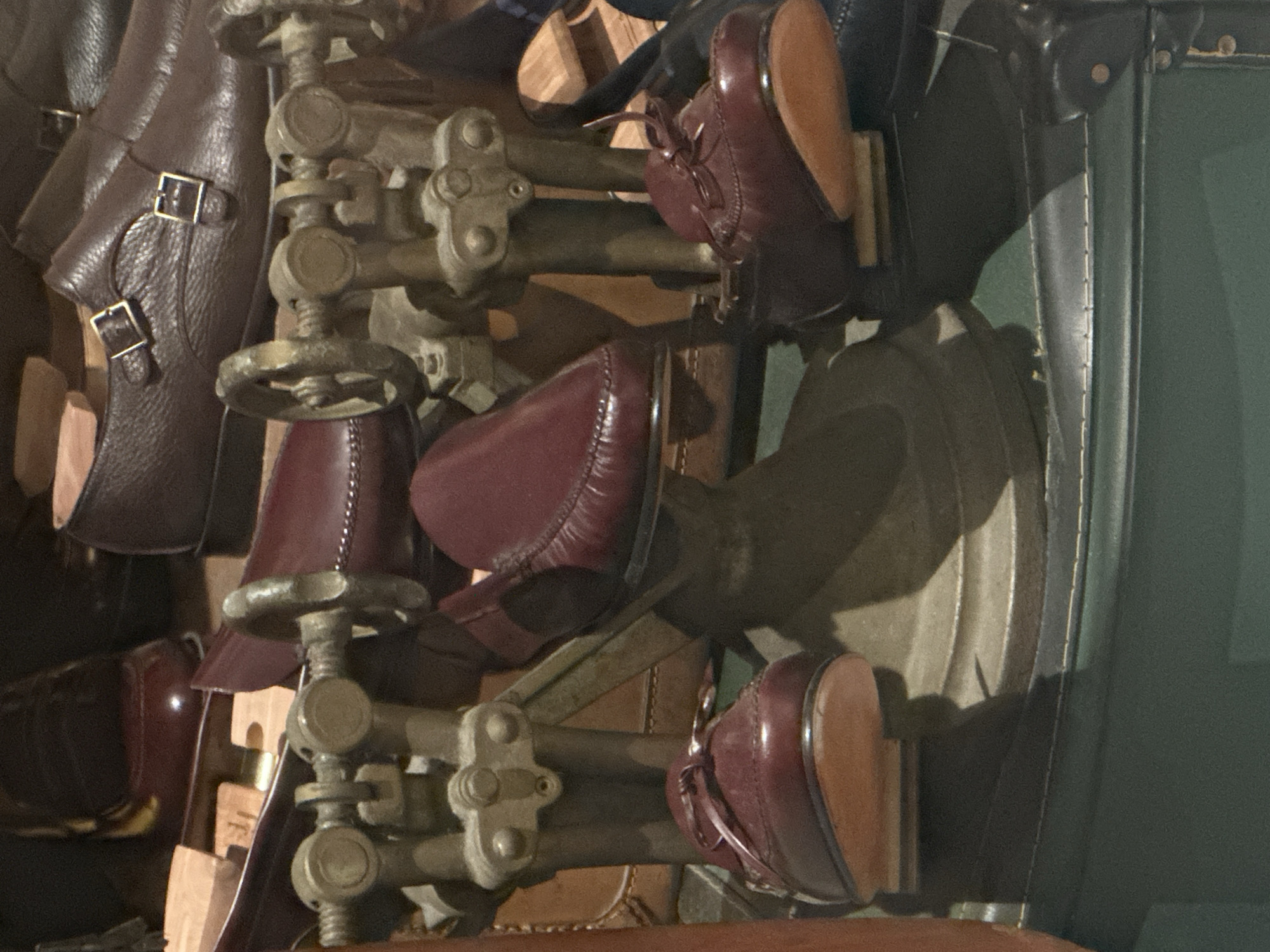 The image captures a close-up, vertically oriented view of a collection of handcrafted leather shoes and intricate metal shoe lasts, suggesting a shoemaker's workshop or a display in a bespoke shoe store, potentially located in Taormina, Italy.
The primary subjects are several pairs of polished leather shoes in rich dark brown and burgundy hues. Some shoes feature double brass buckles, indicative of monk strap styles, while others appear to be laced moccasins or boat shoes with prominent stitching. Adjacent to and supporting these shoes are elaborate, vintage-looking metal shoe lasts or stretchers. These mechanisms are made of an aged, brass-colored metal, featuring gears, screws, and hand-turn wheels, designed for shaping or maintaining footwear. The texture of the metal suggests durability and fine engineering.
Other dark brown leather items, possibly components of shoes or small bags, are visible on the left side, also adorned with similar brass buckles. Portions of light-colored wooden shoe trees or display stands are also interspersed among the leather goods.
The background is dark and somewhat indistinct, with deep green and brown tones, suggesting an indoor setting with subdued lighting. Shadows play across the surfaces, highlighting the textures of the leather and the metallic components, adding depth and a sense of craftsmanship to the scene. The items are arranged in a way that suggests a vertical display or storage system, possibly a wall-mounted rack showcasing the artisan's tools and products. No people are visible, and there is no discernible text in the image. The overall impression is one of traditional artisanship, quality materials, and a dedication to the craft of shoemaking.