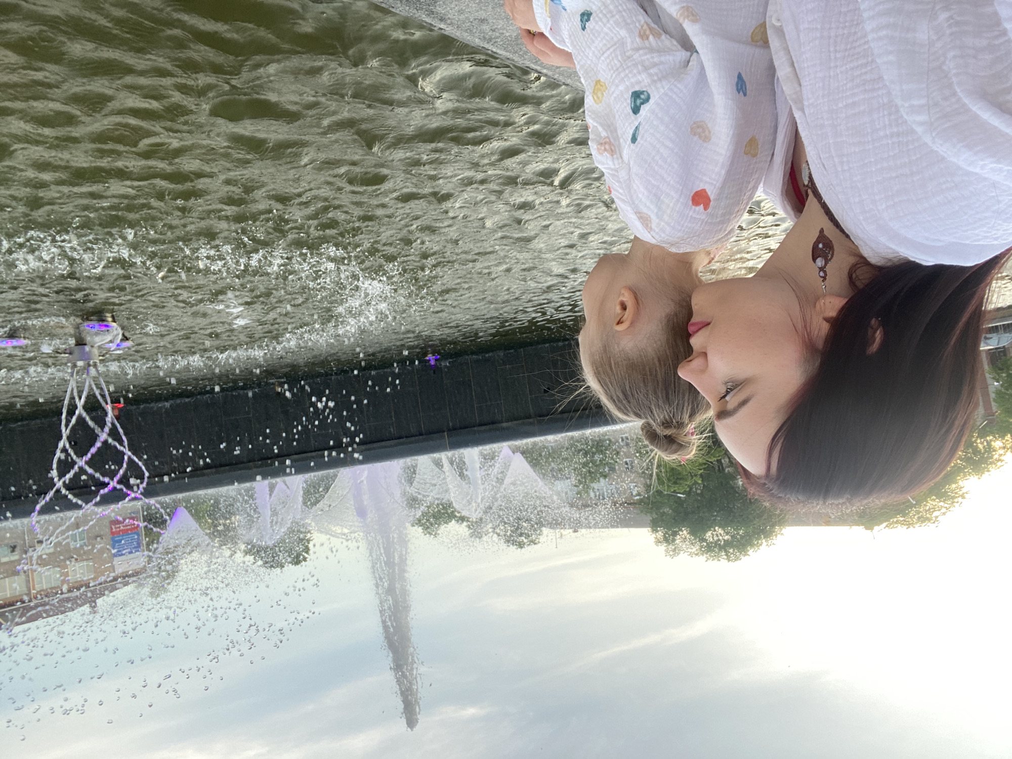 A woman and a small child are positioned next to a fountain in Chaikovsky, Russia. The woman has dark hair and is wearing an earring. The child has fair hair pulled into a bun and is wearing a white garment with small, multicolored heart patterns. Both figures are close to the edge of the fountain. The fountain features a central vertical jet of water as well as illuminated decorative features including a net-like structure. The water has a dark, reflective surface. Buildings and trees are visible in the background.