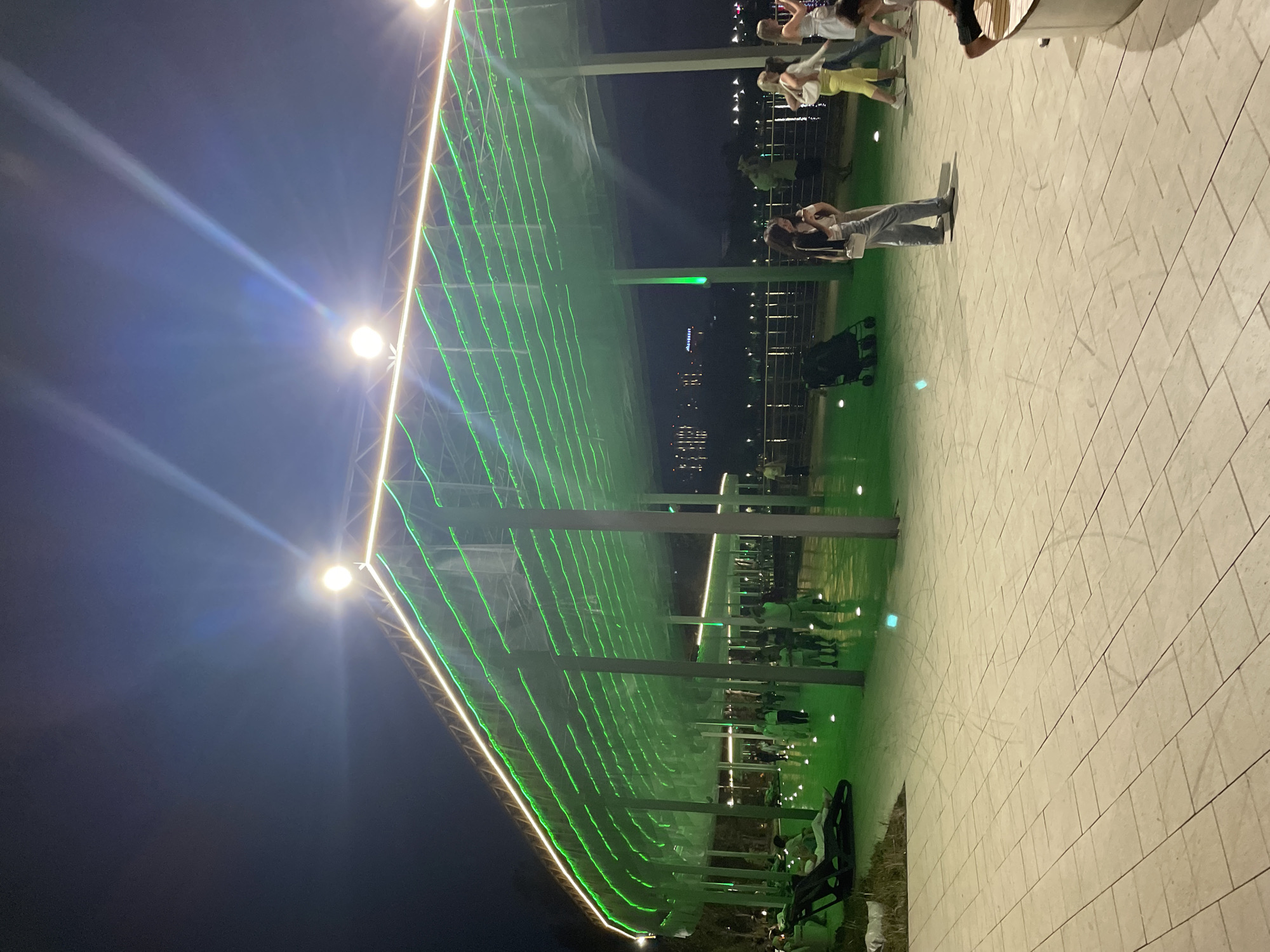 The image depicts an outdoor area at night, likely a park or promenade, illuminated by vibrant green and white lights. The scene features a large, curved canopy structure supported by columns, adorned with strings of green lights. A white light strip runs along the top edge of the canopy. The ground is paved with light-colored square tiles, and there are small circular lights embedded in the ground, casting a green glow.
Several people are visible in the image, engaged in various activities. In the foreground, on the left, a person lies on a lounge chair. Further back, groups of people are walking, some appear to be socializing, and a child's stroller is parked. A woman in the mid-ground is walking towards the right, wearing a white top and grey pants. In the far right of the image, a person is sitting on a curved bench.
In the background, a body of water is visible, with lights from buildings and possibly a shoreline reflected on its surface. The sky is dark, indicating it is nighttime. The overall atmosphere is one of leisure and public gathering.
This scene is characteristic of the waterfront area of Almaty, Kazakhstan, specifically the areas around Kok-Tobe hill or the Central Park, which often feature modern architectural designs with decorative lighting for evening enjoyment.