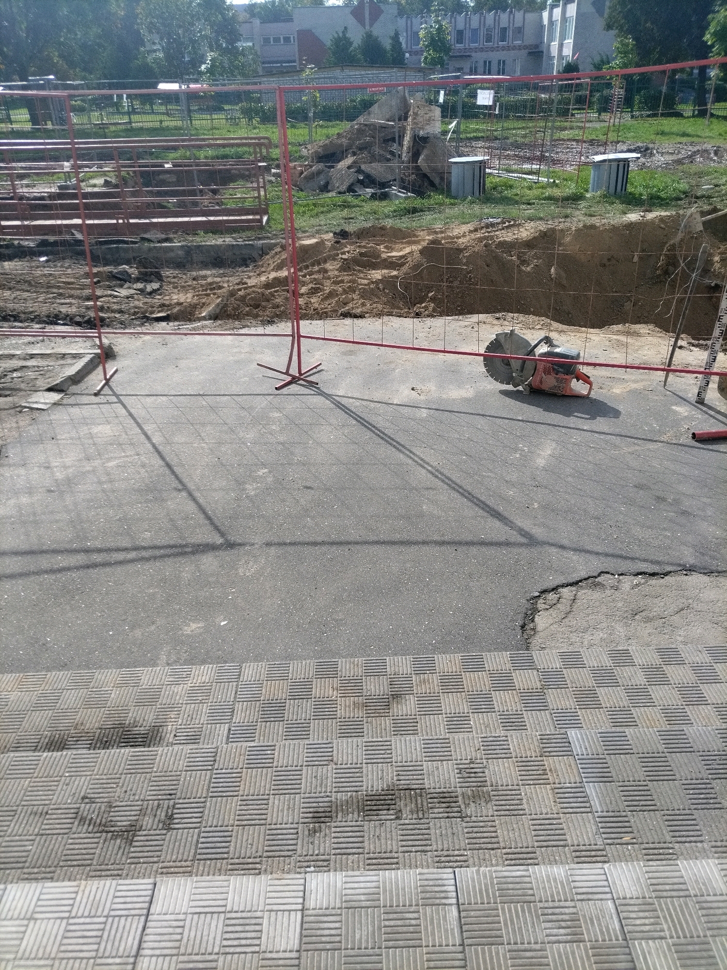 The image shows a construction site with a red metal fence. In the foreground, there is a patterned paved area. Behind this, a section of asphalt is visible, with a cut-off saw on the right side, resting on the asphalt. Beyond the asphalt, there is a large pile of dirt and debris, with more red fencing. In the background, there are trees and buildings, some of which appear to be residential or educational institutions. The scene suggests ongoing construction or repair work in a city environment.