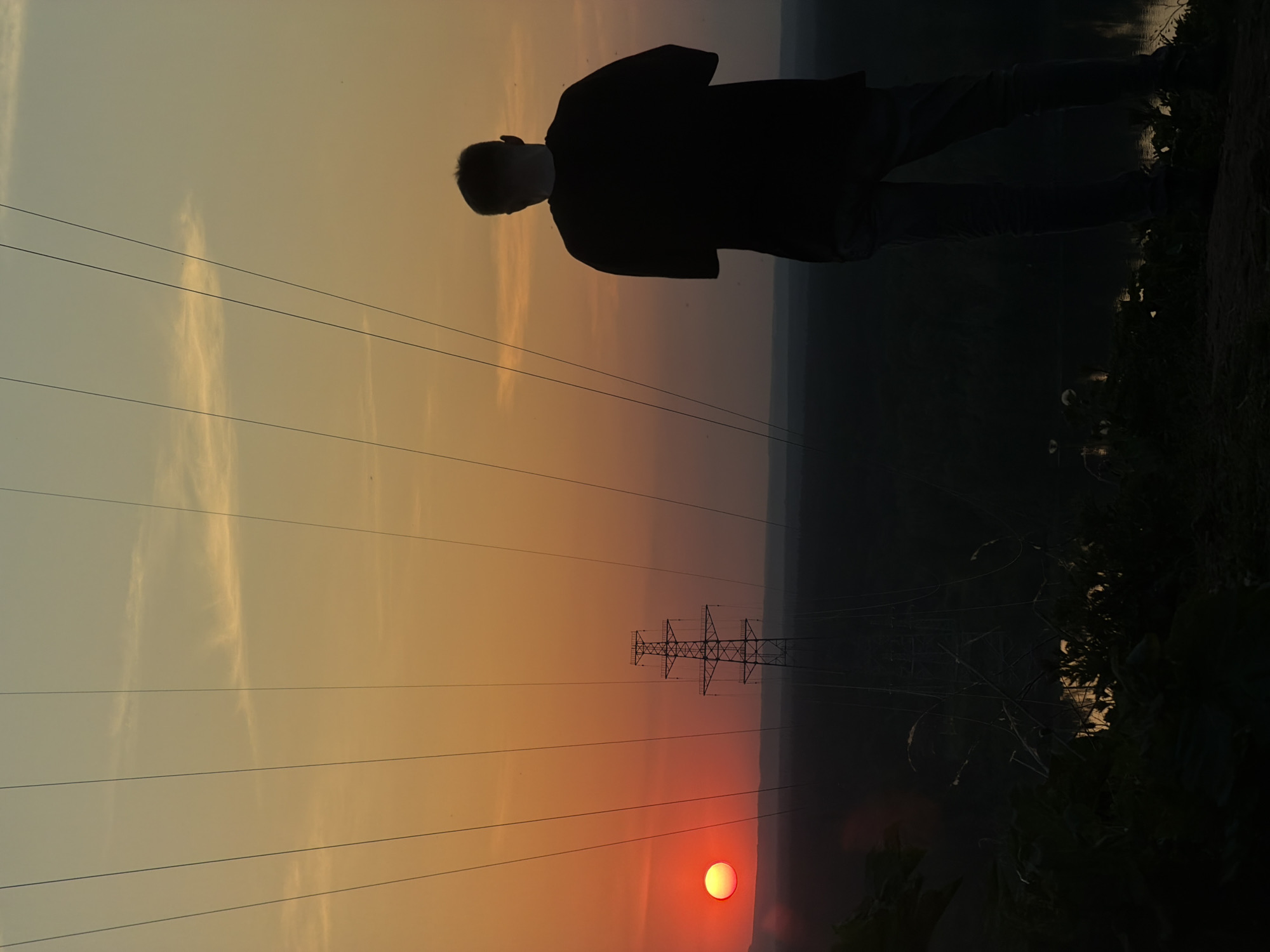 The image depicts a silhouette of a person standing on a raised area, possibly an embankment, overlooking a landscape in Noviy, Russia at sunset. The sun is low on the horizon, appearing as a bright red orb. Power lines stretch across the sky, and a utility pole is visible in the distance. The environment is predominantly dark with a range of foliage, suggesting the presence of vegetation.