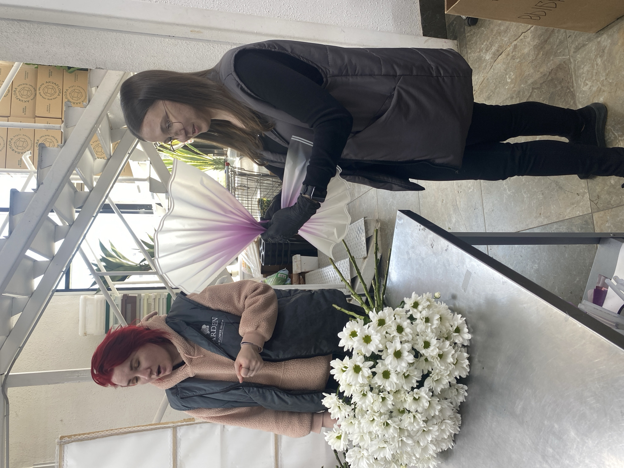 Two women are arranging a bouquet of white daisies. One woman, with long brown hair and glasses, is wearing a black top, black pants, black shoes, and a gray vest. She is holding the wrapped bouquet in her gloved hands. The other woman has red hair, wears a pink jacket under a gray vest, and stands next to the daisies. They are working at a metal table. The location appears to be a florist shop in Yekaterinburg, Russia, as evidenced by the inscription “GARDEN FLOWER BOUTIQUE” on the vest of the red-haired woman and the Russian text on boxes in the background.