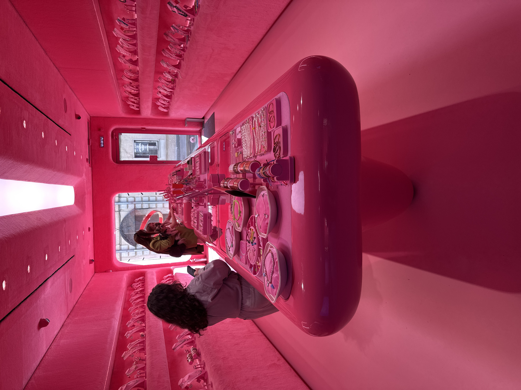 This media file captures the intensely monochromatic pink interior of what appears to be a retail store or boutique, located in Moscow, Russia. The entire space is saturated with various shades of pink, from deep fuchsia to softer rose.
The main subjects are two women and the store's elaborate pink decor. In the foreground, a woman with dark, curly hair is seen from behind, wearing a light-colored jacket or shirt. She is leaning over a long, highly polished, rounded pink counter, appearing to examine the merchandise displayed there. Further back, another woman, wearing a green top, is visible near a large window or glass door.
The dominant feature is the extensive use of pink. The walls and ceiling are covered in a textured, possibly felt or velvet, pink material. Along one side, recessed circular lights illuminate the wall, and a long, narrow light strip runs horizontally above. The central, elongated counter is a smooth, reflective pink, showcasing numerous items such as bracelets, necklaces, and earrings arranged in small pink and white dishes and stands. Similar items are displayed on pink-covered ledges or shelves along the walls.
The activity taking place is shopping or browsing. The women are engaging with the displayed goods. Through the large window at the far end of the counter, an urban street scene is visible, with reflections of buildings and structures, indicating it is daytime.
Notable details include the striking all-pink aesthetic, creating a distinct and immersive environment. A red sign with white Cyrillic text reading "ВЫХОД" (EXIT) is visible above what appears to be a door frame near the window. The overall impression is one of a modern, stylish, and highly themed retail space.