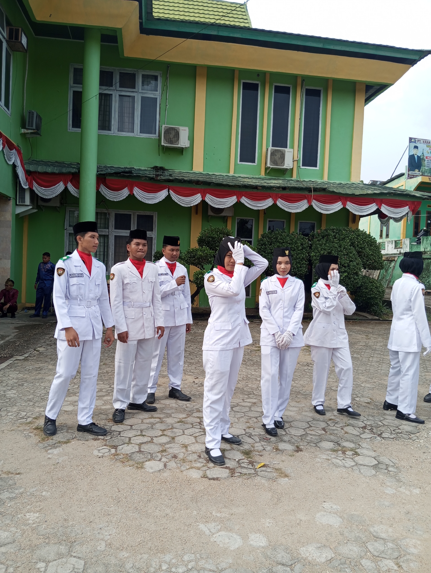 In front of a two-story green building with yellow accents and a green tiled roof in Pematang Reba, Indonesia, seven individuals in white uniforms with red neck scarves and black caps stand on a paved area. The building has several windows and air conditioning units visible. Red and white bunting hangs above the first-floor windows. Two individuals in blue attire and one person in red attire are partially visible in the background to the left. A sign is mounted on the right side of the building. The individuals in white uniforms are facing towards the left of the frame, with some touching their faces or adjusting their caps. The paved area is a mix of hexagonal and irregularly shaped stones.