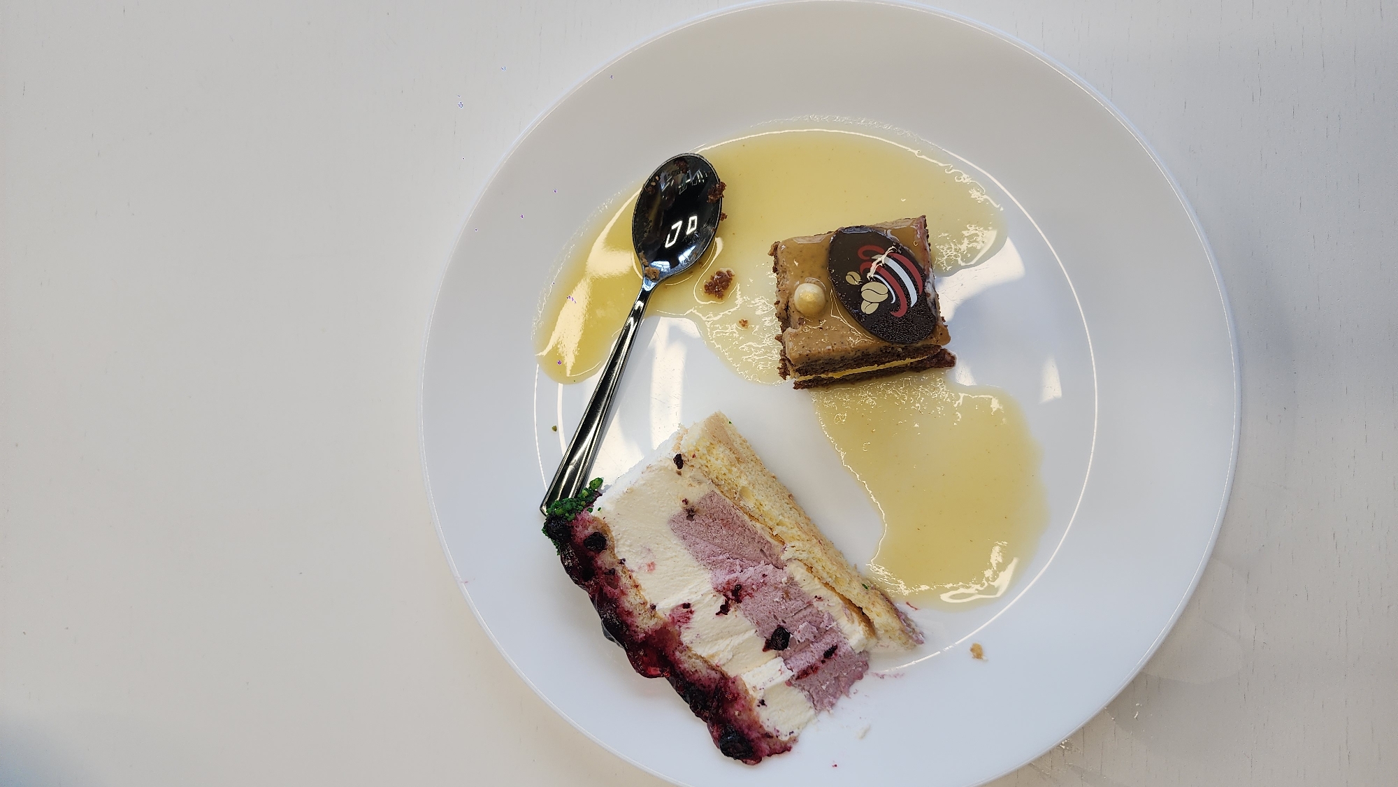 A white plate contains two pieces of cake, a dessert sauce, and a spoon. One cake piece is a square, topped with a chocolate decoration and nuts. The other piece is a slice with layers of cake and cream, with a dark berry coating on one side. The sauce is a translucent, pale yellow color. The spoon is silver and partially covered in the sauce. The background is a white surface. The image does not contain information about Moscow, Russia, or people.