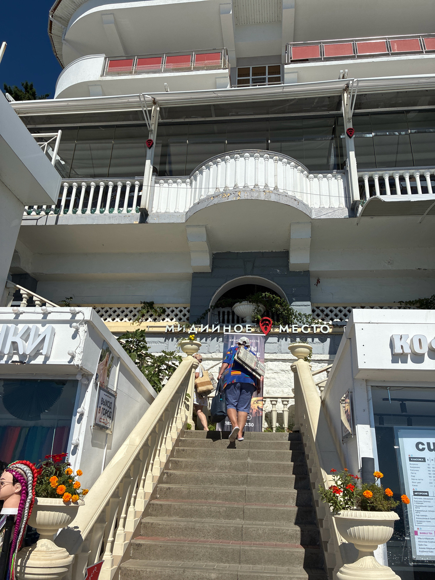 The image shows a white, multi-story building with a curved facade and red balconies. A wide, stone staircase with a white balustrade leads up to the building. Two people are ascending the stairs. The person in front is wearing a blue and yellow striped shirt and blue shorts, carrying a large white object over their shoulder. The person behind them is wearing a white top and light-colored skirt, carrying a bag. Several shops are visible on either side of the staircase. One sign above the shops reads "МИЛЛИОННОЕ МЕСТО" (Millionth Place) in Russian, with a red location pin icon. Another sign on the left reads "КИ" in large white letters. The scene appears to be outdoors on a sunny day, with a clear blue sky. The building's architecture suggests it might be a hotel or a resort. Based on the Cyrillic signage, the location is likely in Russia. The style of the building, particularly the curved balconies and ornate balustrades, is characteristic of Art Deco or Stalinist Empire style architecture, which is present in Sochi, Russia, a city known for its post-Soviet resort architecture.