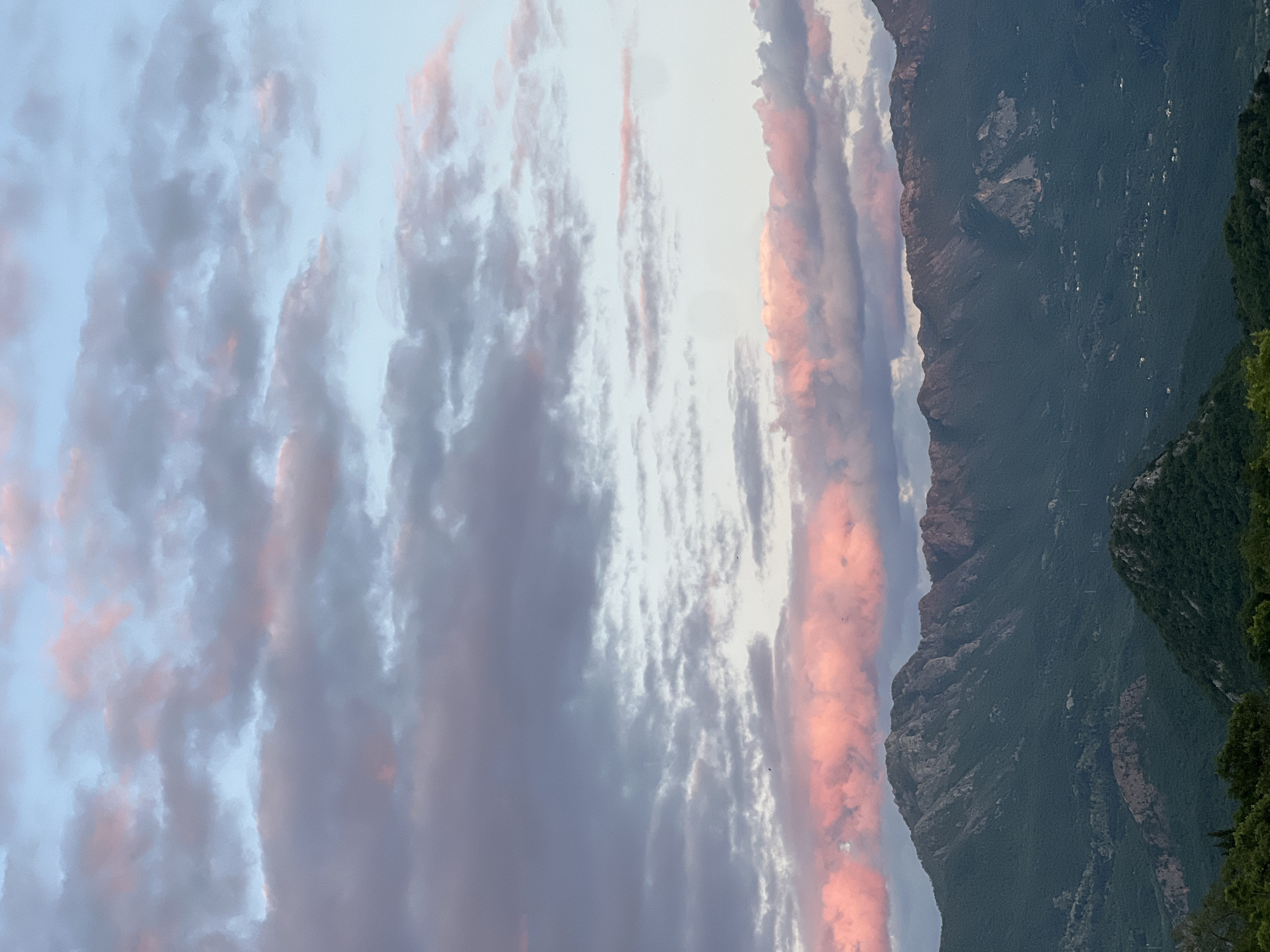 The image depicts a landscape in Sotonići, Montenegro, featuring a mountain range partially obscured by clouds. The sky is filled with clouds that are tinged with pink and gray hues. The mountain range is dark green with some visible rock faces. The foreground shows partial tree foliage. The image is oriented vertically.