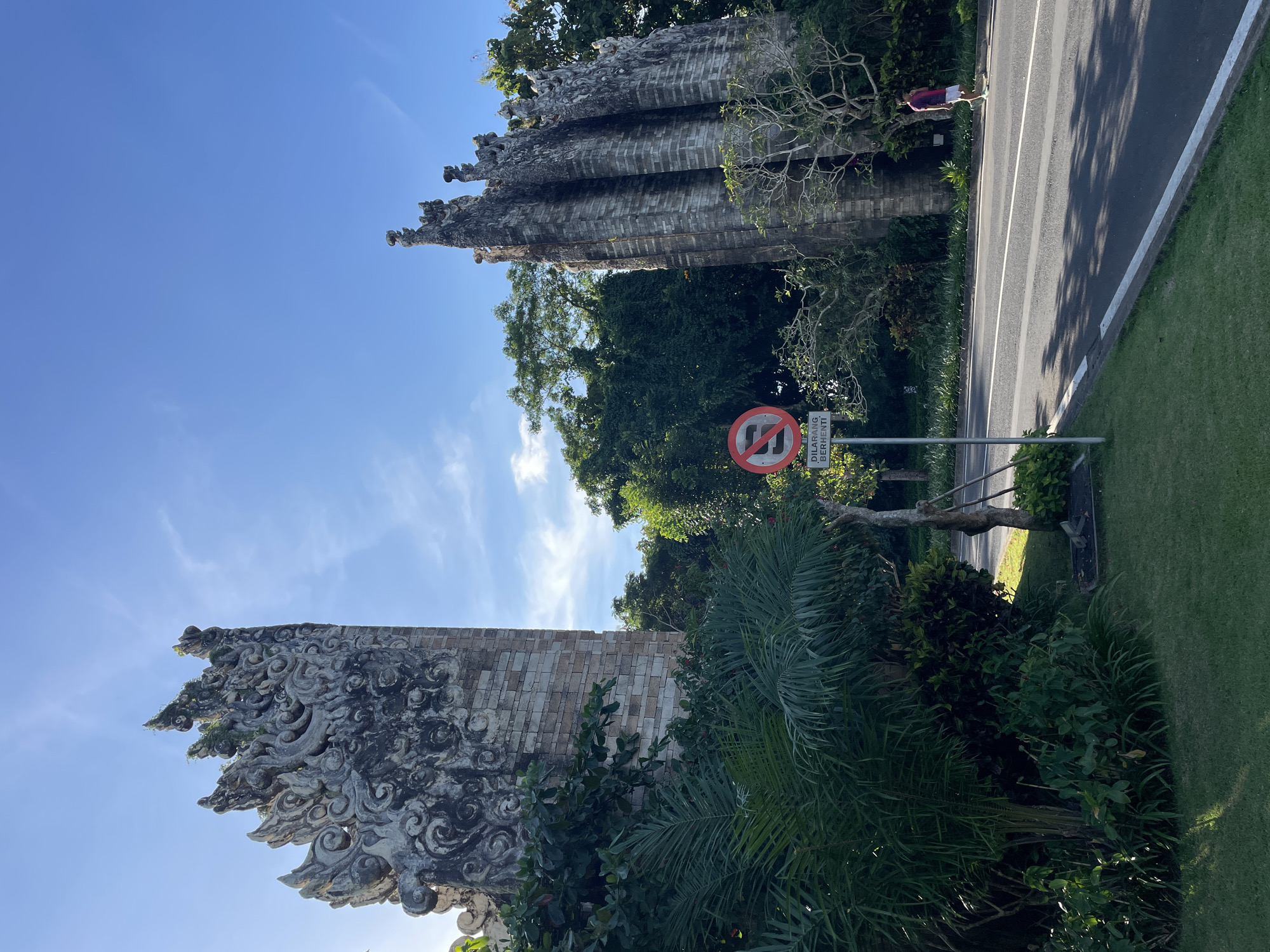 The image depicts a road flanked by elaborate stone structures, likely gates, in Kuta Selatan, Indonesia. The structures are intricately carved with traditional Balinese designs. A 'No Stopping' road sign is visible beside the road. A person is walking in the distance on the edge of the road. The environment includes lush tropical vegetation and a clear blue sky with scattered clouds.