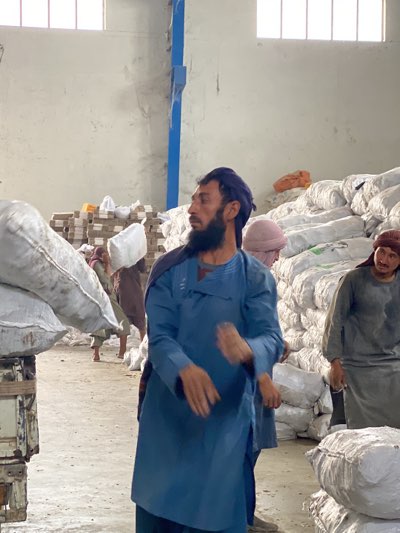 In a warehouse in Shurandam, Afghanistan, several men are working among stacks of white sacks. One man, facing left, wears a blue tunic and a head covering. He has a long beard. Another man stands to the right, wearing a gray tunic and a red head covering. A third man is visible in the background, carrying a white sack on his head. The warehouse has a concrete floor and white walls, with natural light coming in from windows high above. There are also wooden planks and bricks stacked in the background. A blue support beam is visible on the left.