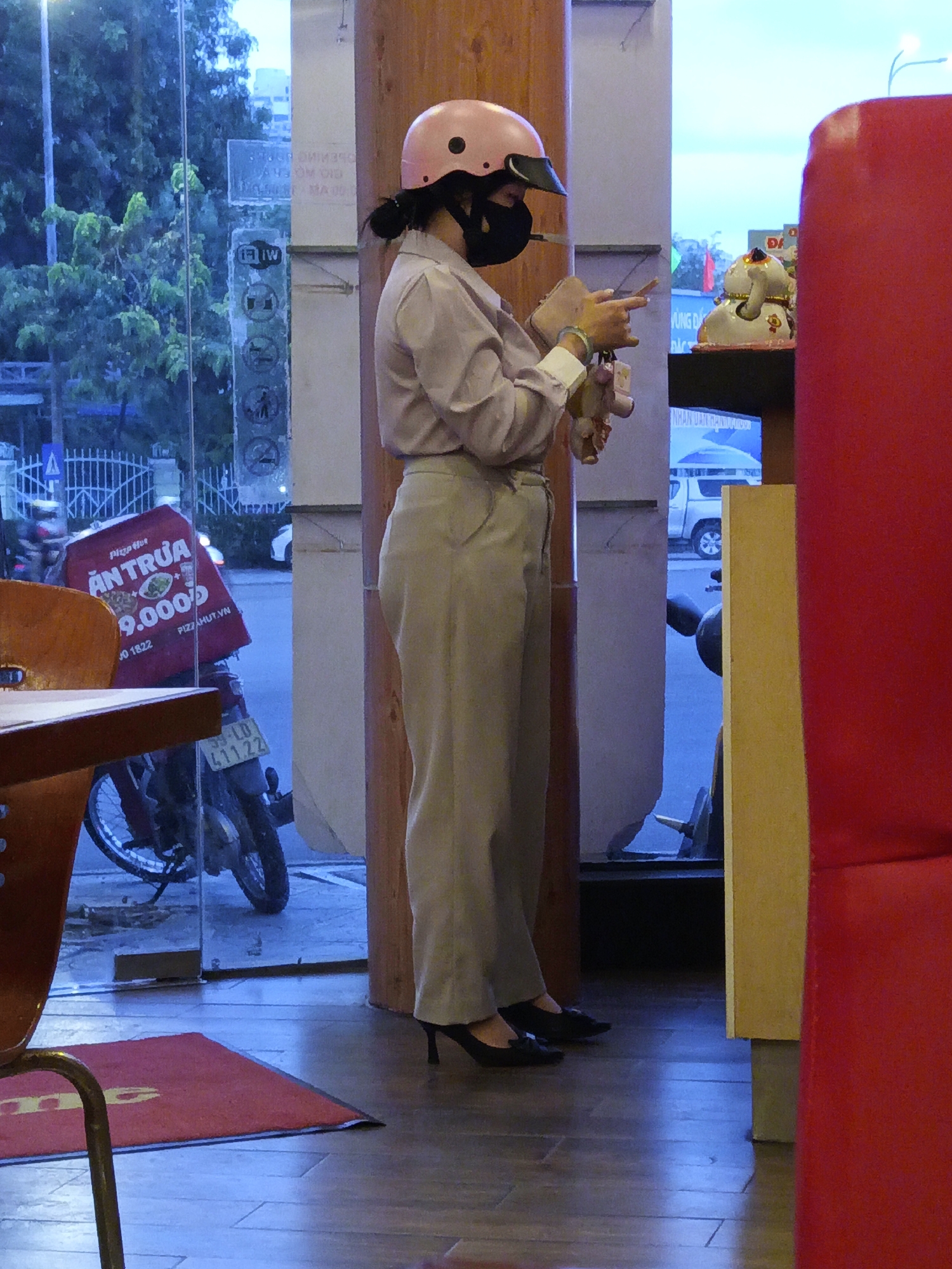 A person wearing a pink helmet and black mask is standing inside a building with a glass window. Outside the window, a motorcycle with a Pizza Hut delivery box is parked. The delivery box is red and displays "AN TRUA 9.000D" and "PIZZAHUT.VN". The motorcycle has a license plate that reads "59-LD 411.22". Inside the building, there are furniture items and a decorative white cat statue. The floor is tiled and reflects light. The scene appears to be in Nha Trang Ward, Vietnam.