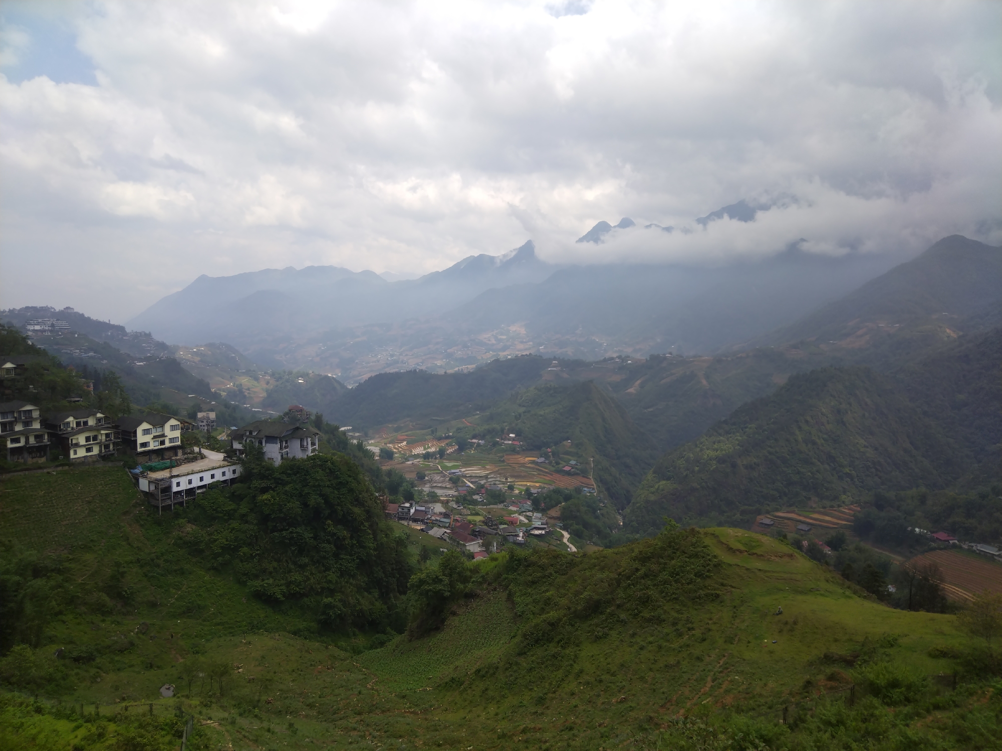 The image presents a landscape view from a high vantage point in what appears to be a mountainous region. In the foreground, there is a green hillside with a visible valley or slope. A group of multi-story buildings, primarily white and grey, are situated on a higher section of the hill on the left side of the image. In the middle ground, a network of hills and valleys stretches into the distance, dotted with what seem to be small settlements or villages. The background features a range of mountains, some partially obscured by clouds or mist. The sky is overcast with a blanket of grey clouds. The overall environment suggests a rural, possibly Asian, location.