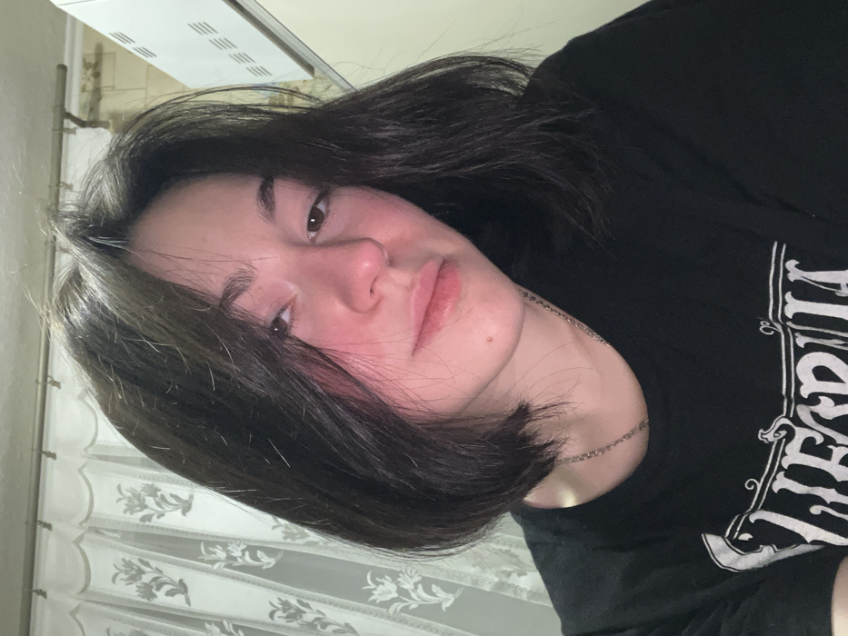 A young woman with dark, short hair, wearing a black graphic tee, is pictured indoors. Her face appears slightly flushed, and she wears a thin silver necklace. She has light skin and dark eyes. A window with patterned curtains is visible in the background, along with a bright, rectangular light fixture on the ceiling. The image is rotated to the right.