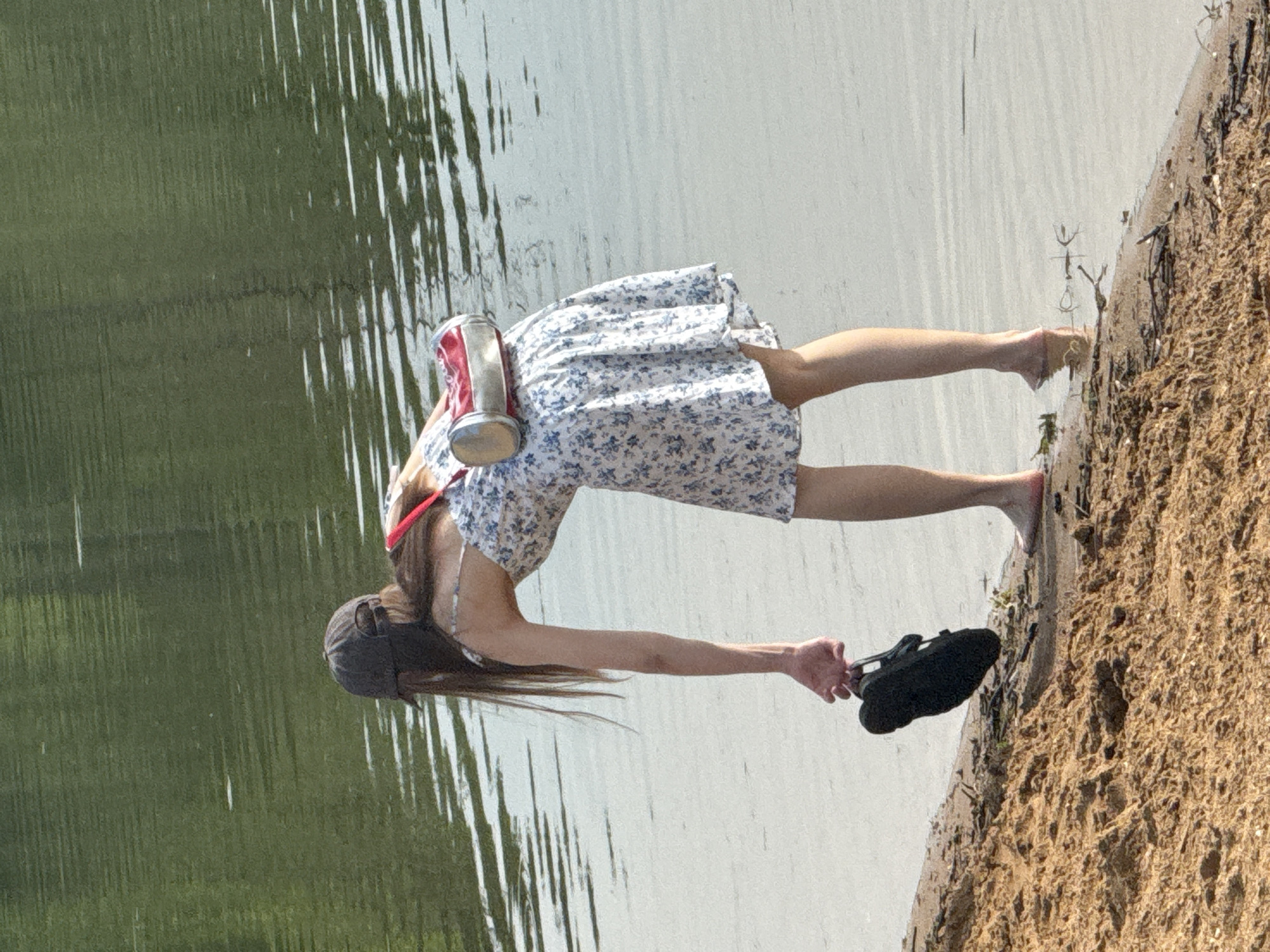 A person stands on a sandy shore near water. The person wears a floral dress, a baseball cap, and carries a black shoe in one hand and a metal container on their back. The water appears calm with tree reflections on the left side. The shore is sandy and uneven with some small plants or debris. The image suggests an outdoor setting by a lake or river. The location, Novosinkovo, Russia, isn't visually verifiable from the image.