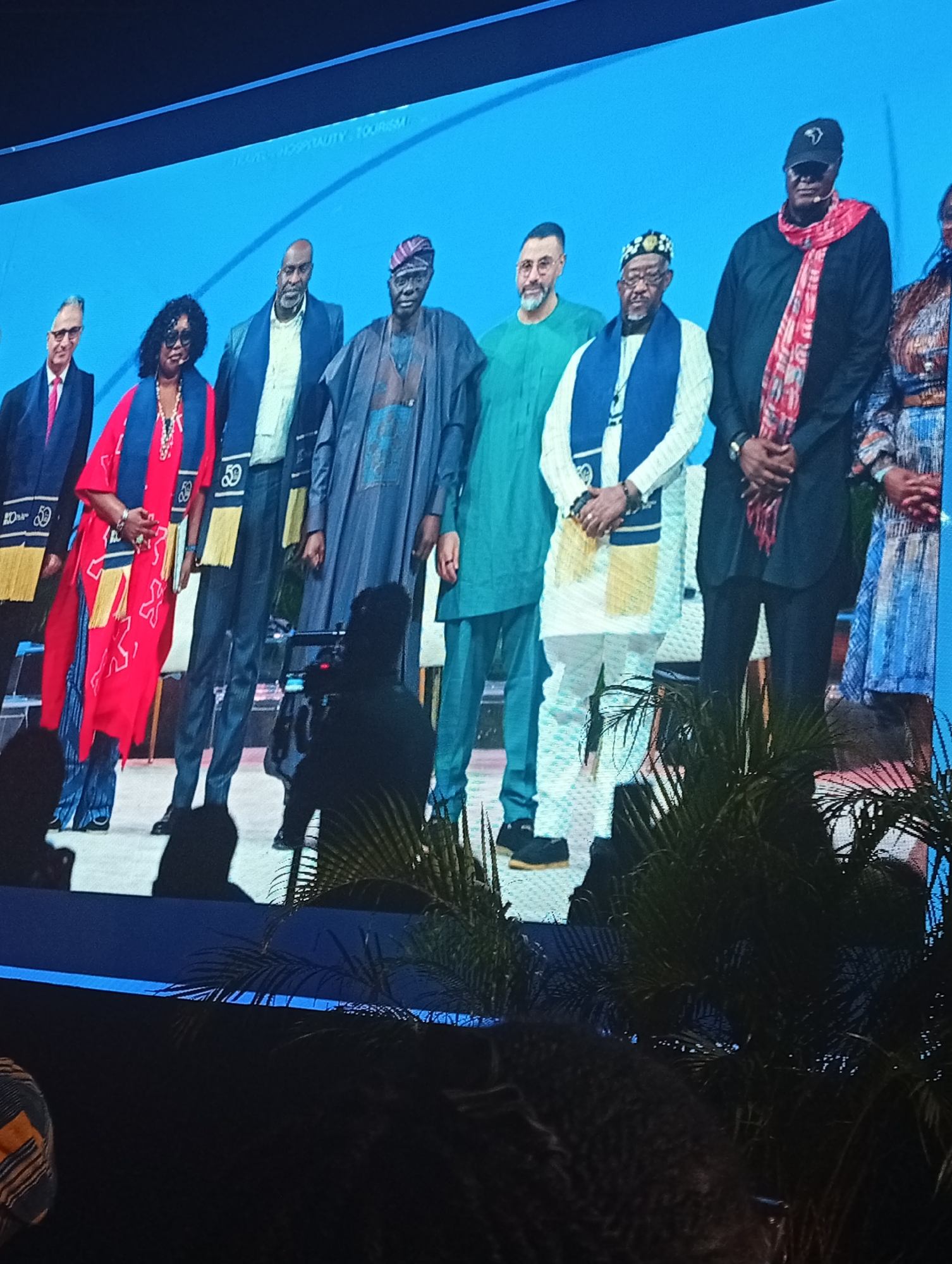 On a brightly lit screen, a group of eight people are standing side by side. They all appear to be of African descent and are dressed in formal attire. Some wear stoles with the number "50" printed on them. The background is a bright blue screen with white text. In the foreground, silhouettes of people and palm fronds suggest the photo was taken from an audience perspective. No city location or evidence of Nigeria, or city of Itirin.