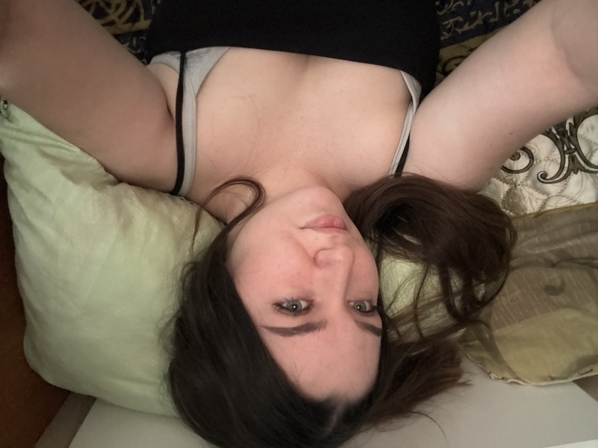 A young woman with dark hair and light skin is lying in bed, looking at the camera. She is wearing a black tank top and a grey bra. Her head is resting on a light green pillow, and there is another pillow with a patterned cover behind her head. The bedding appears to be dark with an ornate pattern. The scene is dimly lit, suggesting it may be nighttime or the room is dark. The location is specified as Borodulino, Russia.